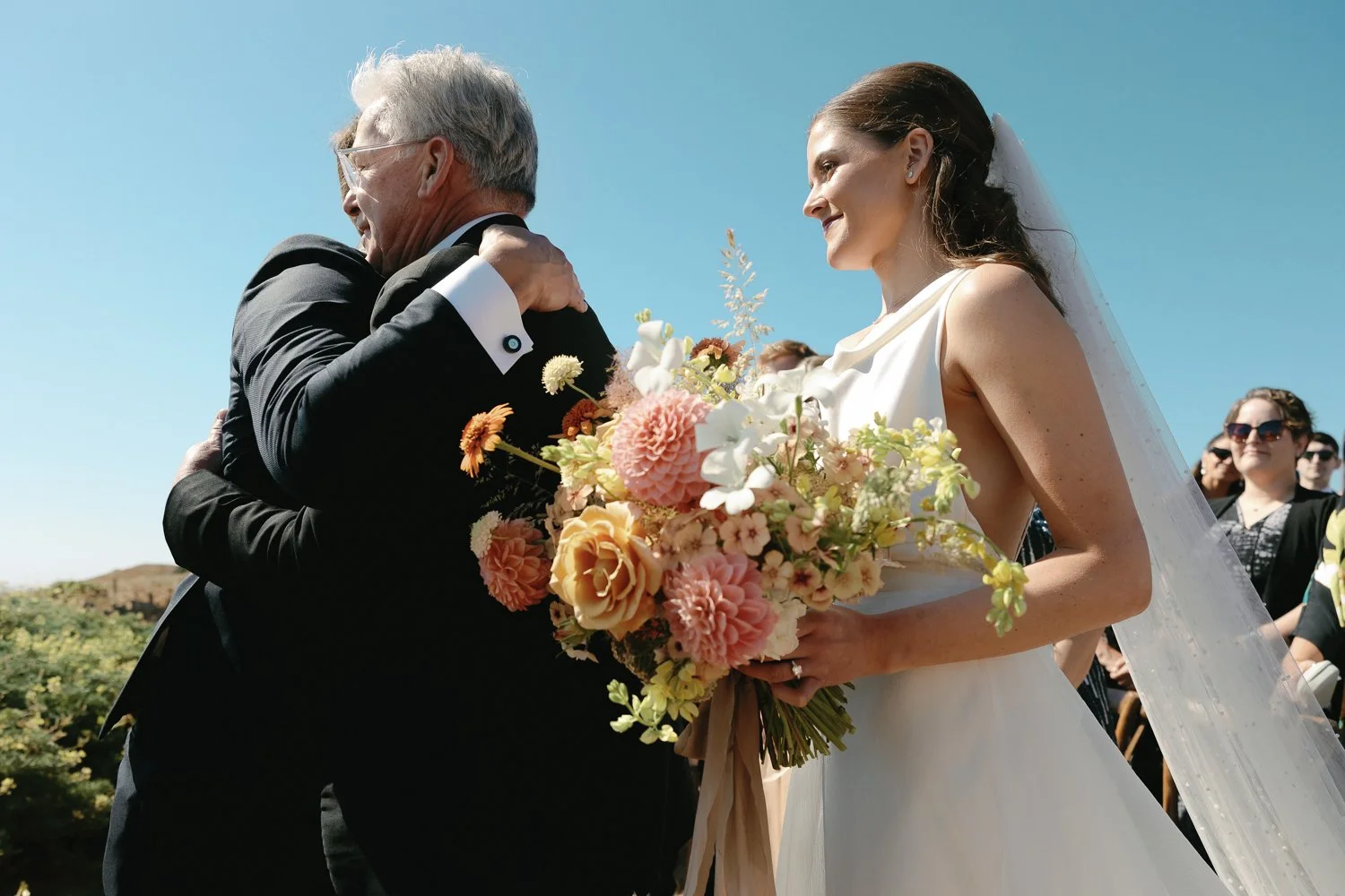 A bride in a white wedding dress holding a bouquet of colorful flowers, hugging an older man in a black suit at an outdoor wedding ceremony under a clear blue sky.
