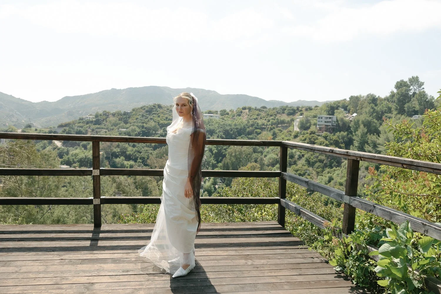 A woman in a white dress and veil standing on a wooden deck with a mountain and green landscape background.
