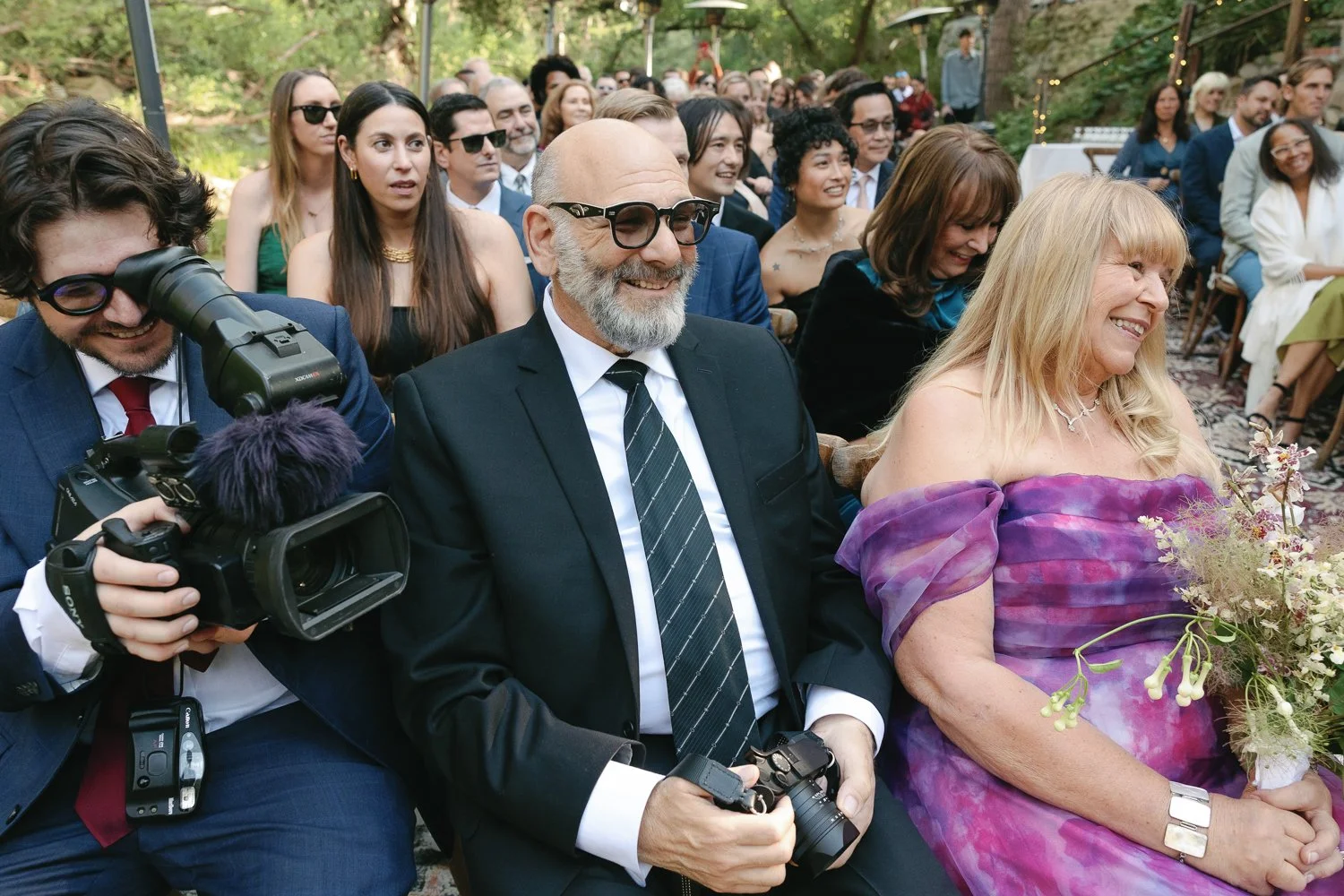 People attending an outdoor wedding, with a focus on a man in a black suit, striped tie, sunglasses, and gray beard, and a woman in a purple off-shoulder dress holding a bouquet of flowers, surrounded by other guests seated on wooden chairs.