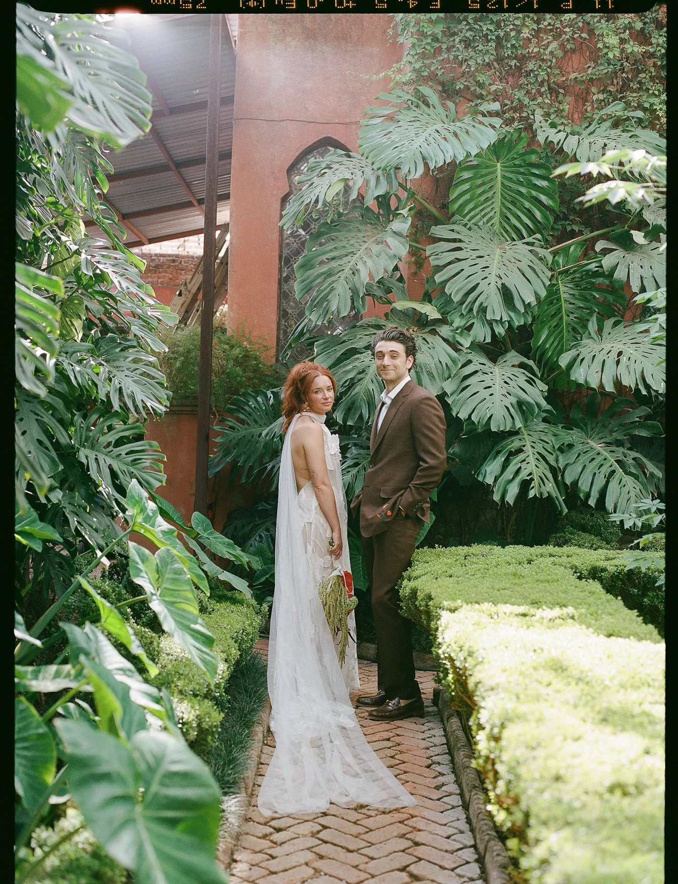 Bride and groom posing on a brick garden path surrounded by lush tropical monstera plants and manicured hedges at casa hyder