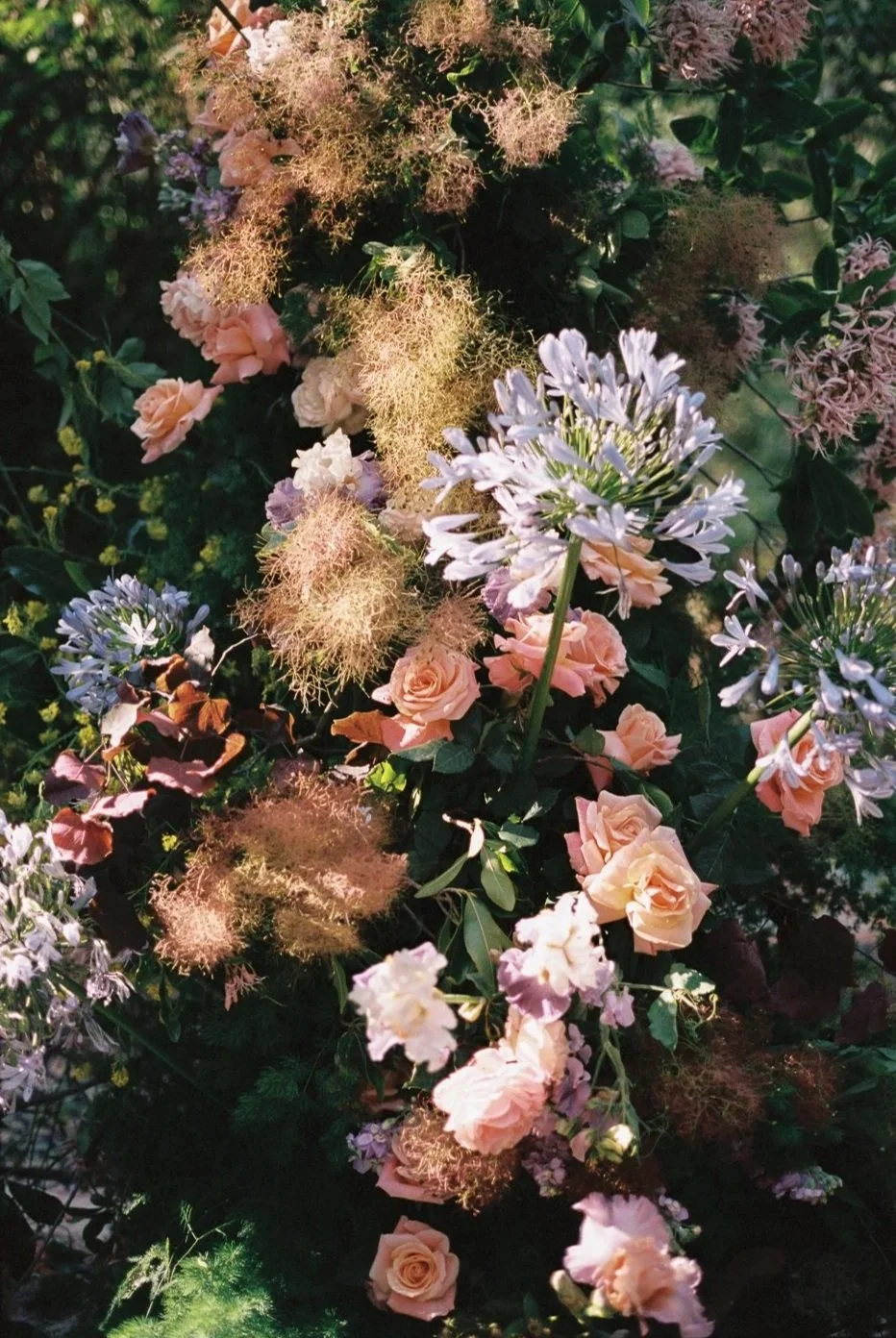A bouquet of various flowers including light purple, pink, and white roses, purple lilies, and fluffy, feathery pink and purple blooms with green leaves.