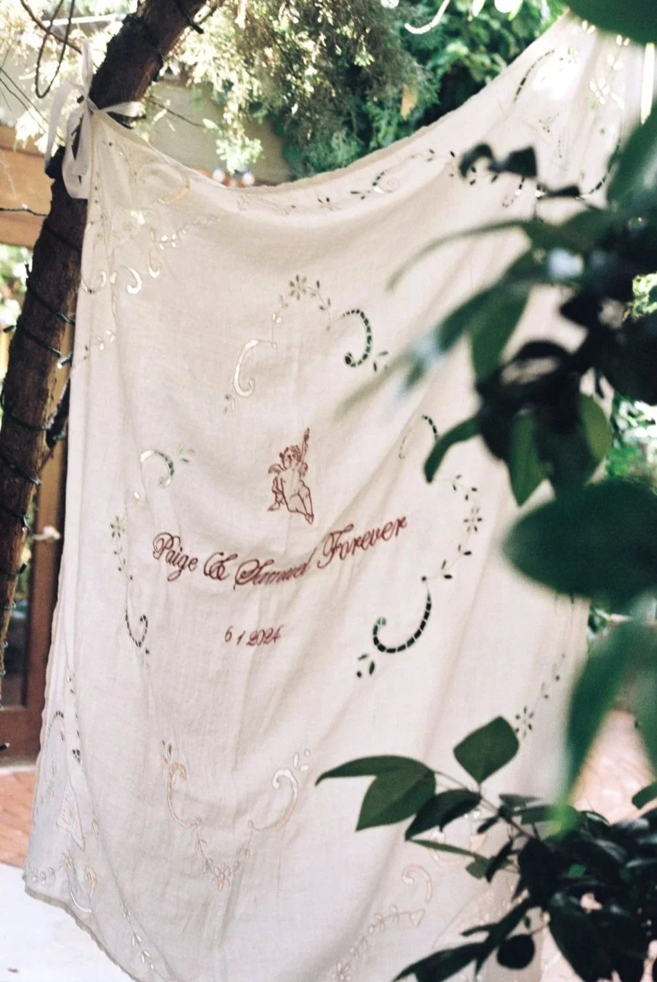 A vintage decorative cloth as a welcome sign with embroidery writing and design hanging from a tree branch, surrounded by greenery. The embroidery reads: 'Paige & Sam Forever 6 1' with a small angel illustration.