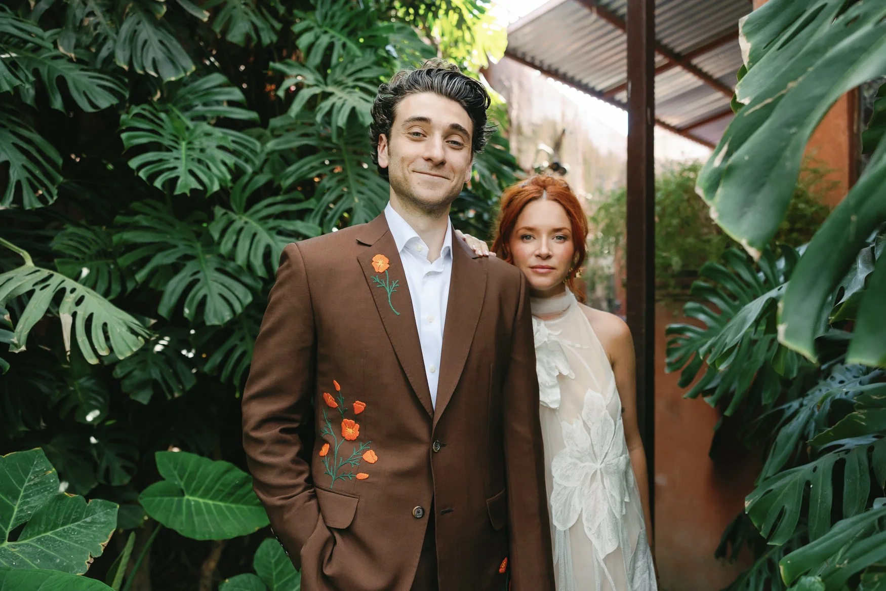Couple posing in the tropical garden at Casa Hyder, wearing a brown floral-embroidered brown suit and white floral applique wedding dress