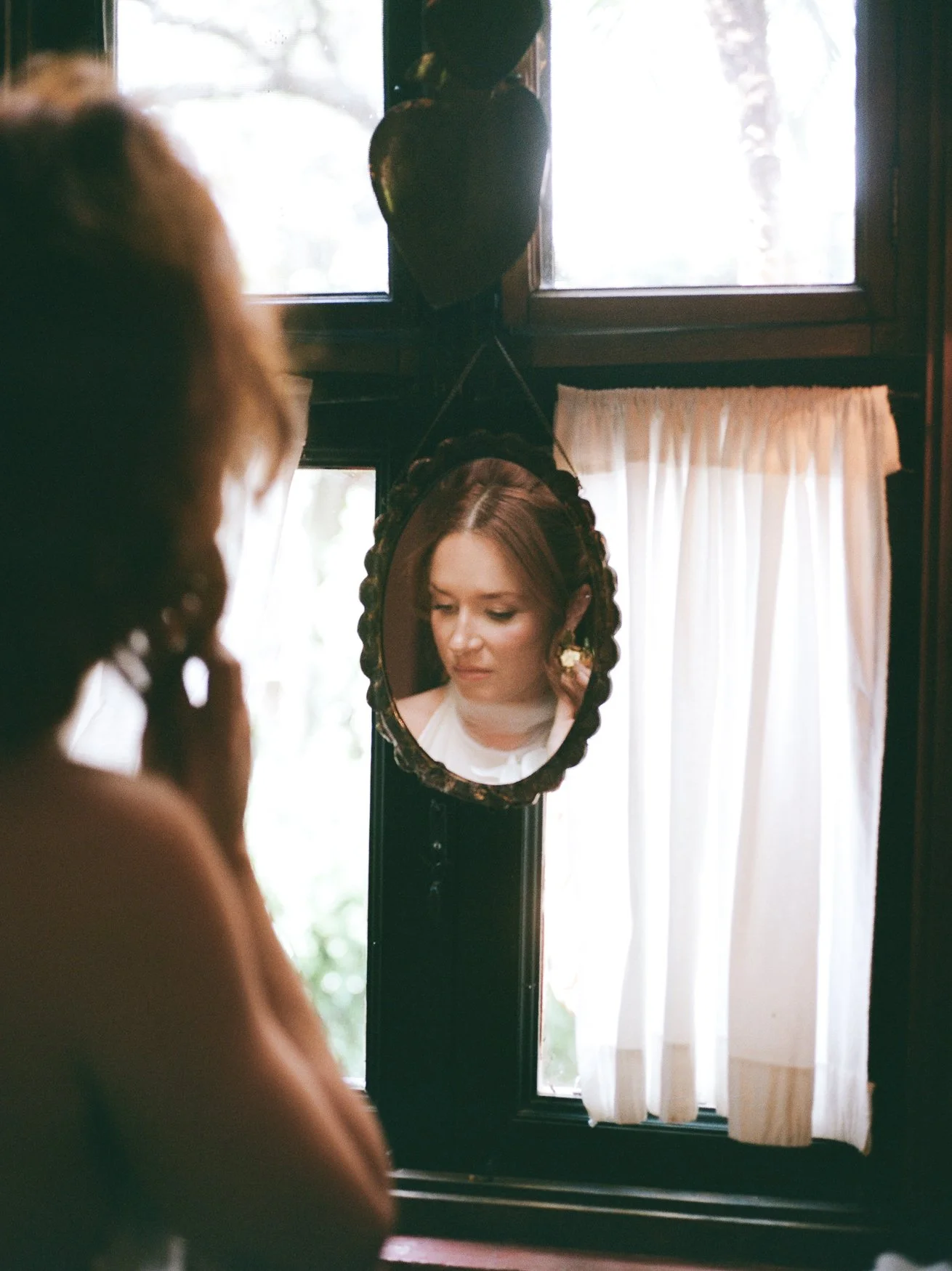 Woman with braided hair and jewelry reflected in an ornate oval mirror beside a sunlit window