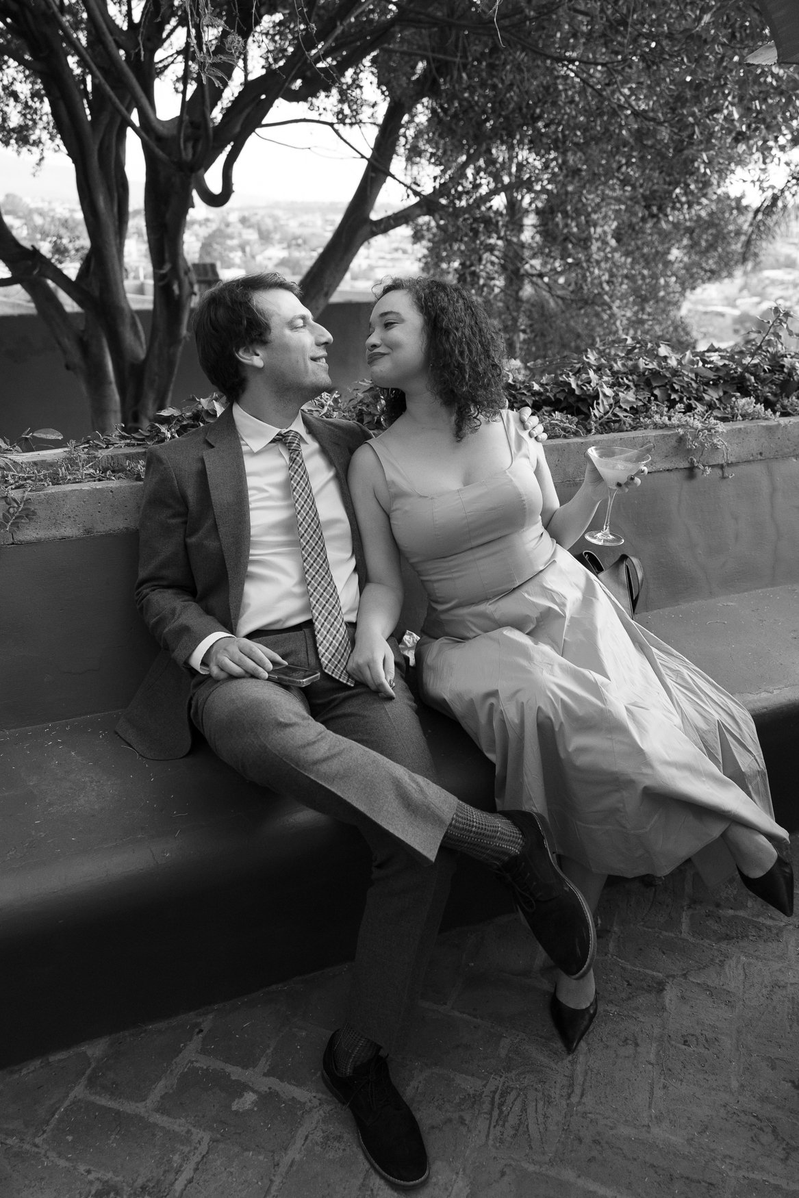 Black-and-white portrait of a well-dressed couple sitting outdoors, gazing at each other as the woman holds a cocktail glass.