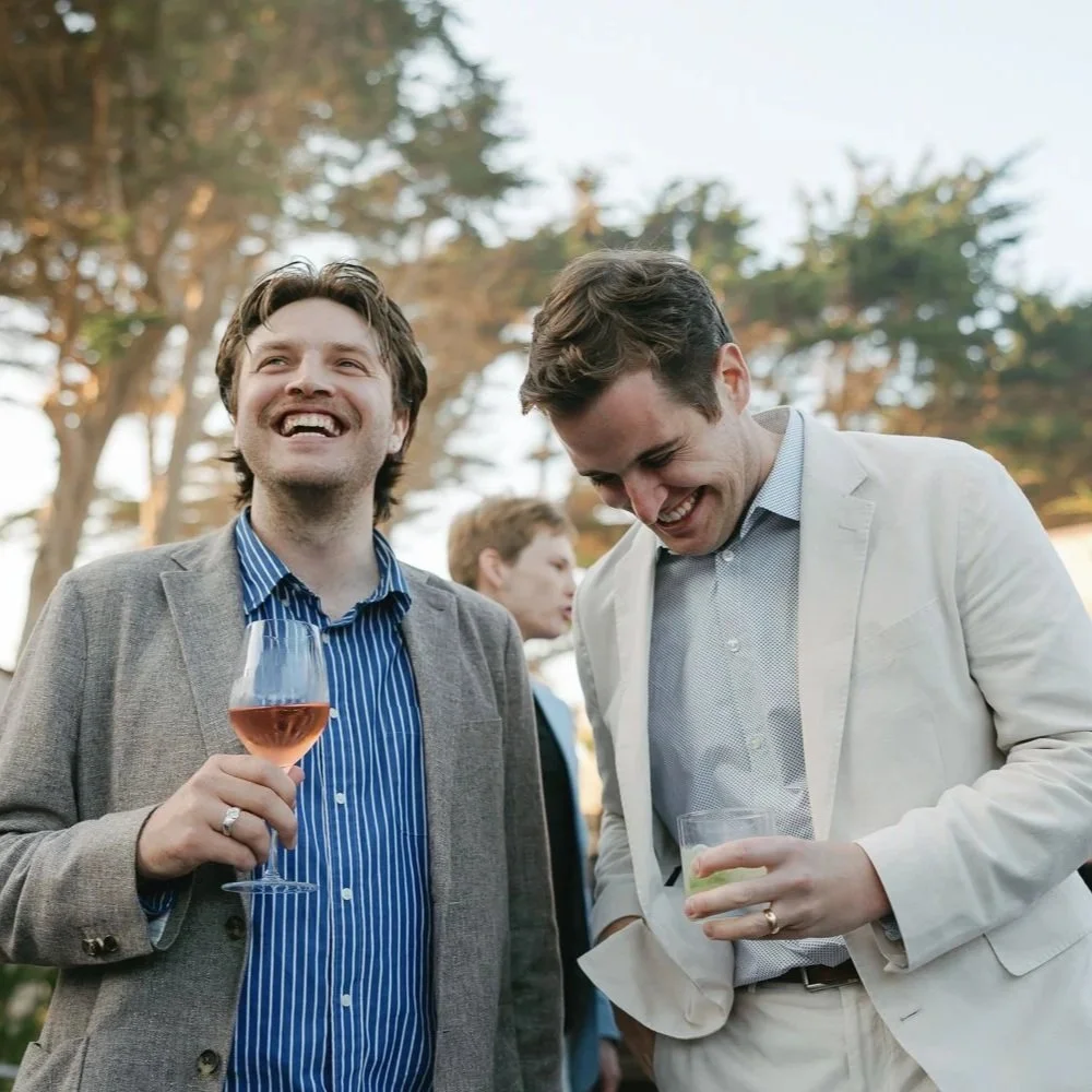 Two men smiling and holding drinks at an outdoor gathering, with trees in the background.