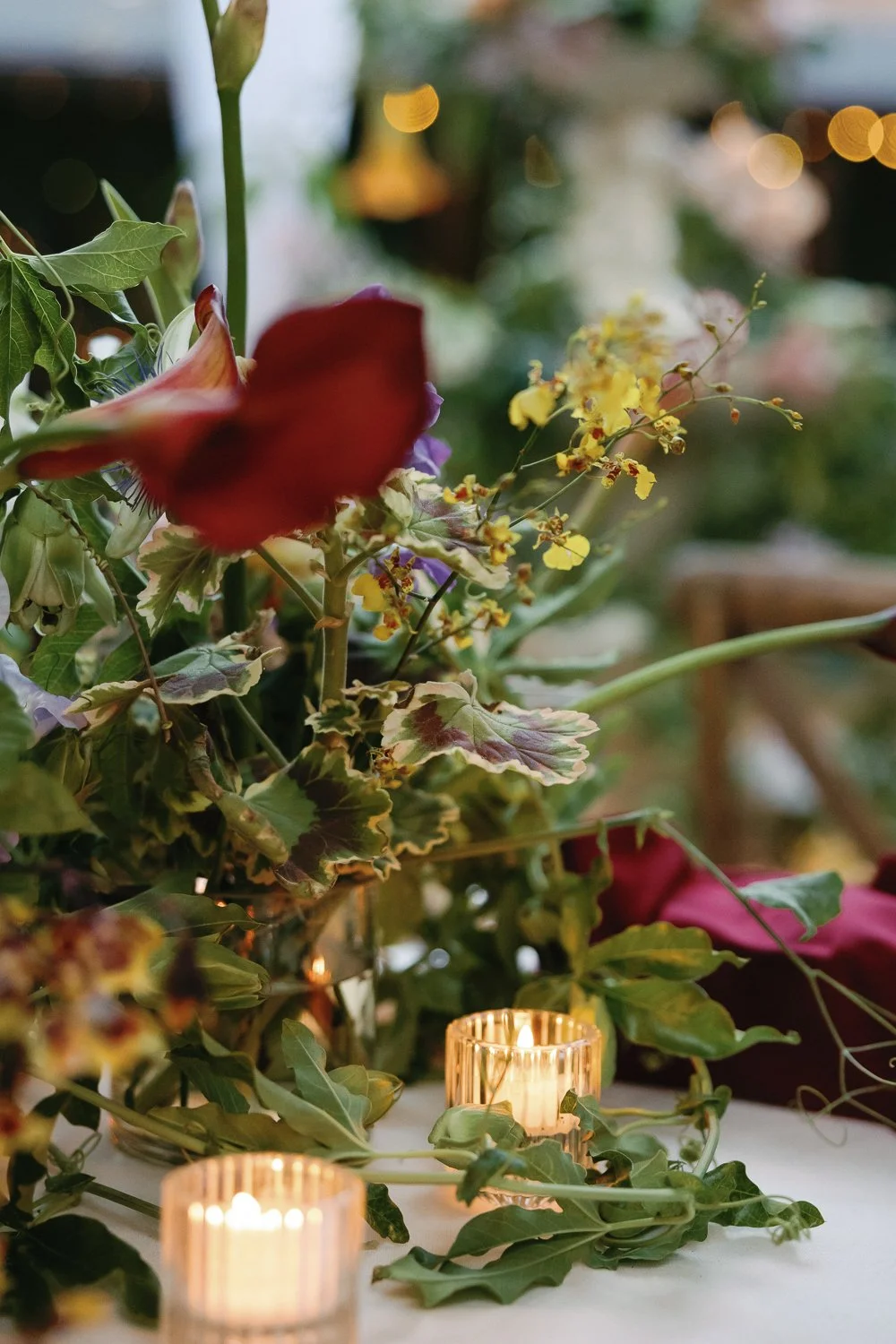 Close-up of a floral arrangement with various green leaves, yellow flowers, and a blurred red flower, with two small lit candles in glass holders in the foreground.
