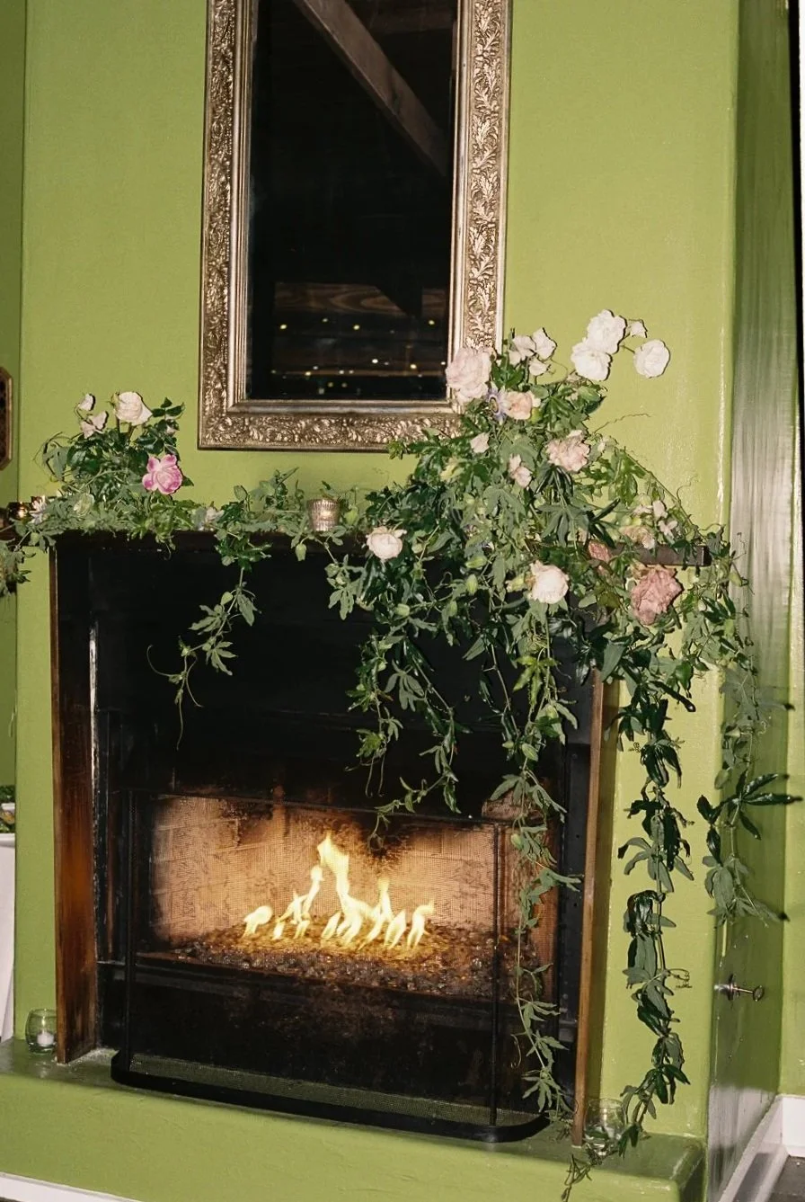 Decorative fireplace with a glowing fire, surrounded by green plants and pink and white flowers on a mantle, with a large ornate mirror above and a green wall.