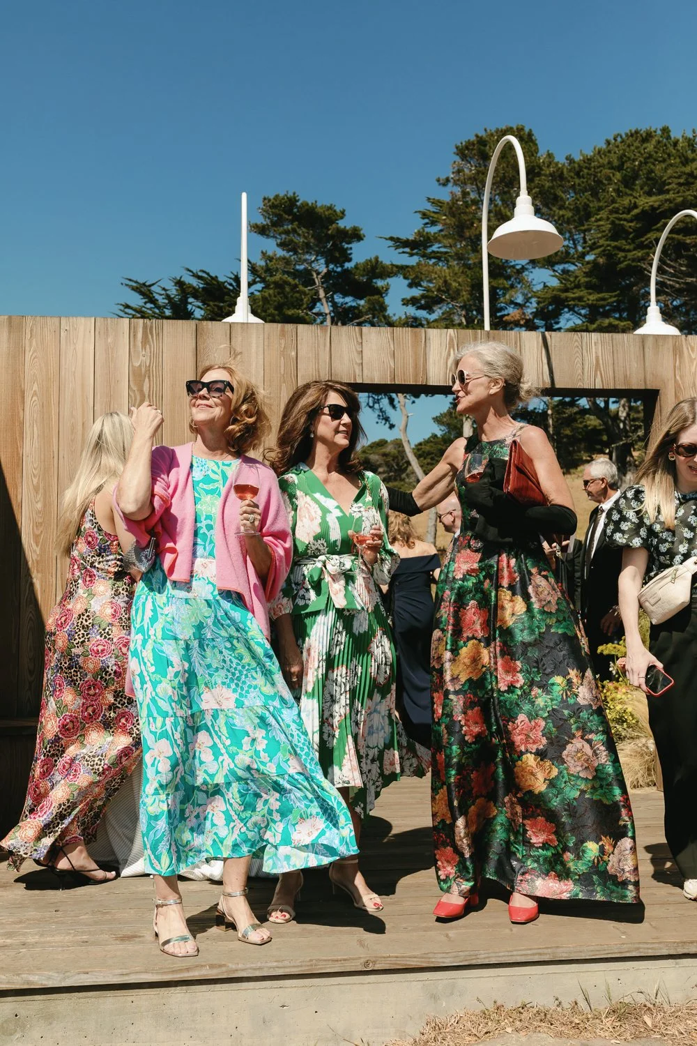 Group of women at an outdoor social gathering, dressed in colorful floral dresses, sunglasses, holding glasses of wine, enjoying sunny weather.
