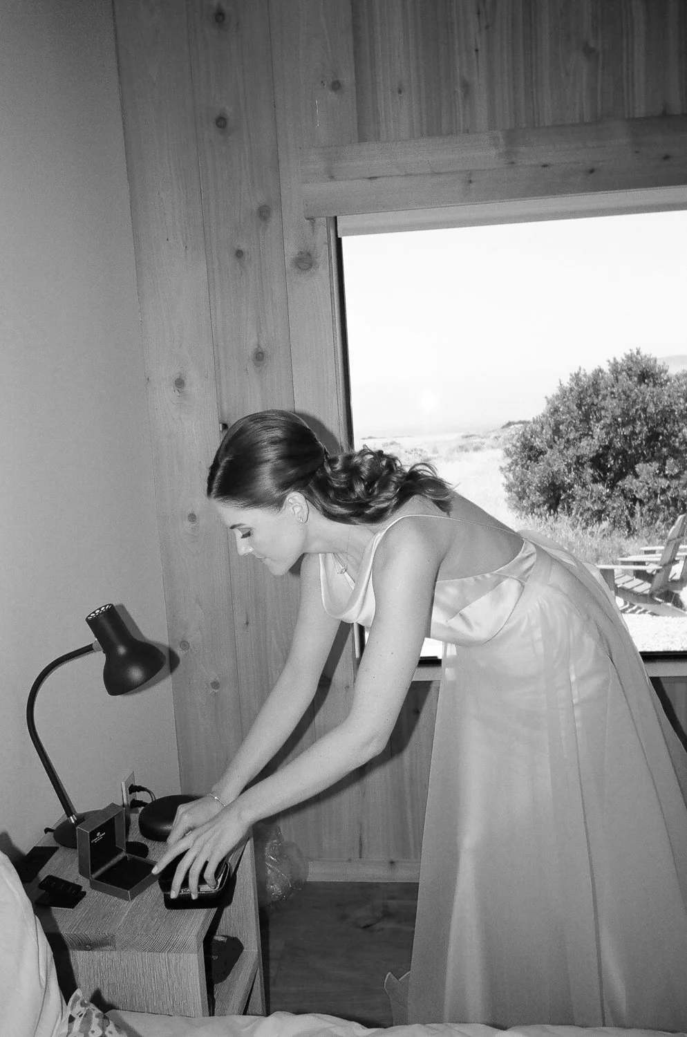 A woman in a satin dress leaning over a bedside table in a rustic room, with a large window showing outdoor trees and landscape.