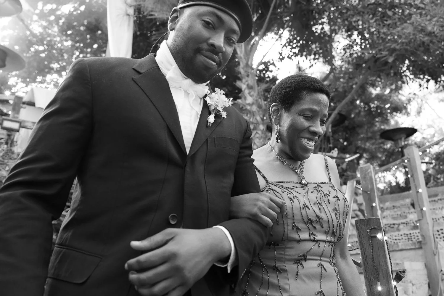 A black-and-white photo of a man and woman smiling and walking outdoors, dressed in formal attire. The man wears a suit with a boutonnière, and the woman wears a detailed dress with jewelry.