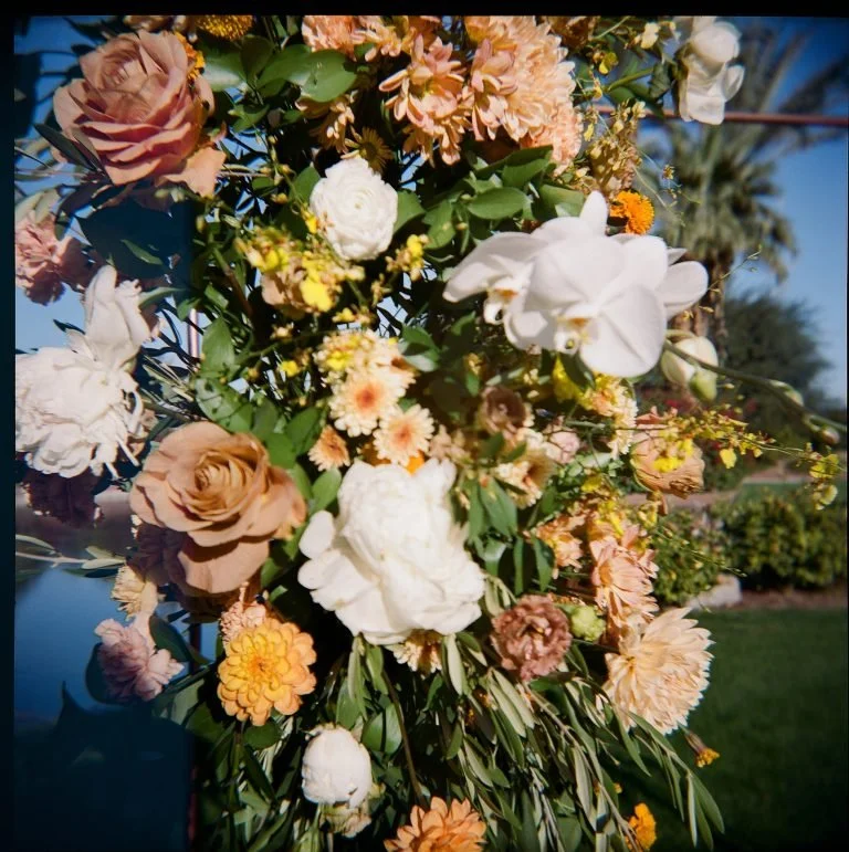 Close-up of a colorful flower arrangement with roses, peonies, and other blooms, with green foliage and a bright blue sky background.