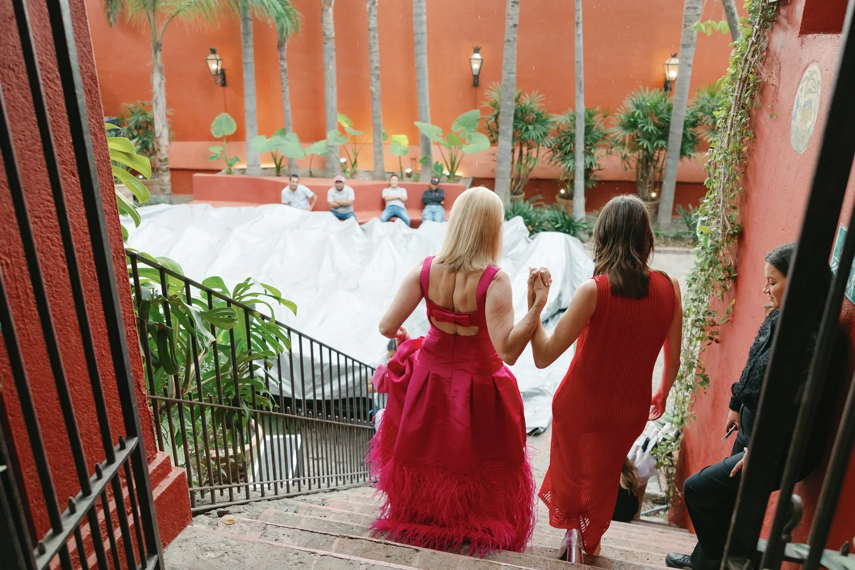 Guests descend a staircase into a tropical courtyard, led by two women in bright pink and red dresses.