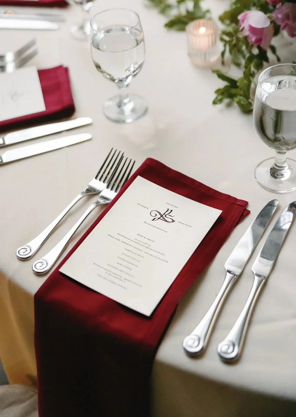 Elegant table setting with a white tablecloth, a red napkin, silverware, a menu, a water glass, and a glass of water, with pink flowers and a candle in the background.