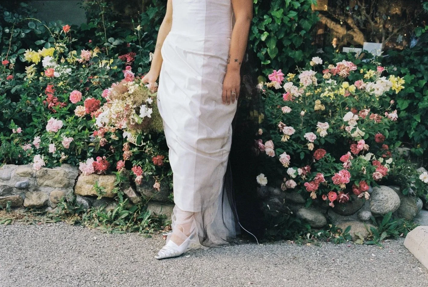 Person in a white dress standing in front of colorful flowers and greenery, holding a bouquet of flowers