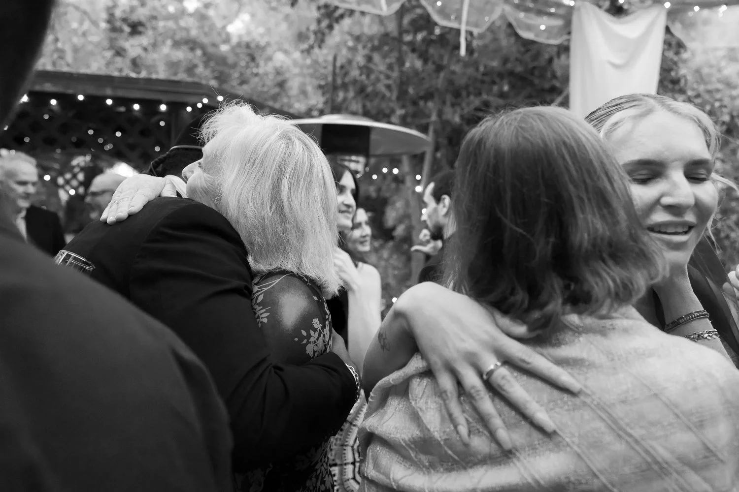 People hugging and smiling at an outdoor gathering or celebration, with trees and lights in the background.