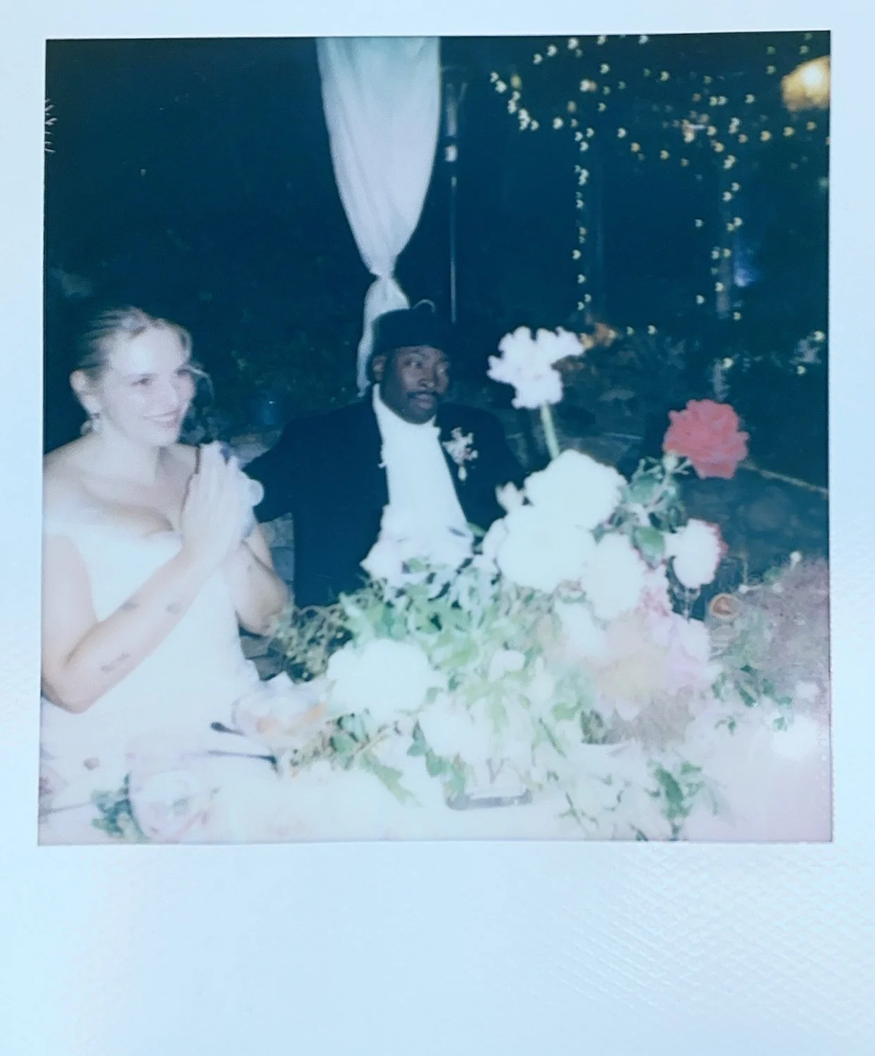 A bride and groom sitting at a table with a large floral centerpiece, celebrating their wedding with string lights in the background.