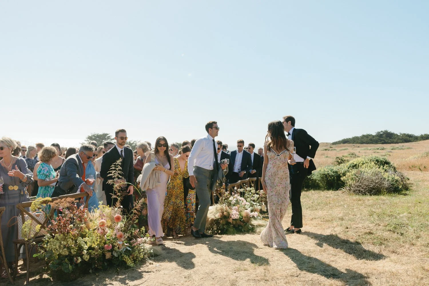 Outdoor wedding ceremony with guests dressed in formal attire, floral decorations, and a scenic landscape with clear blue sky and grassy fields.