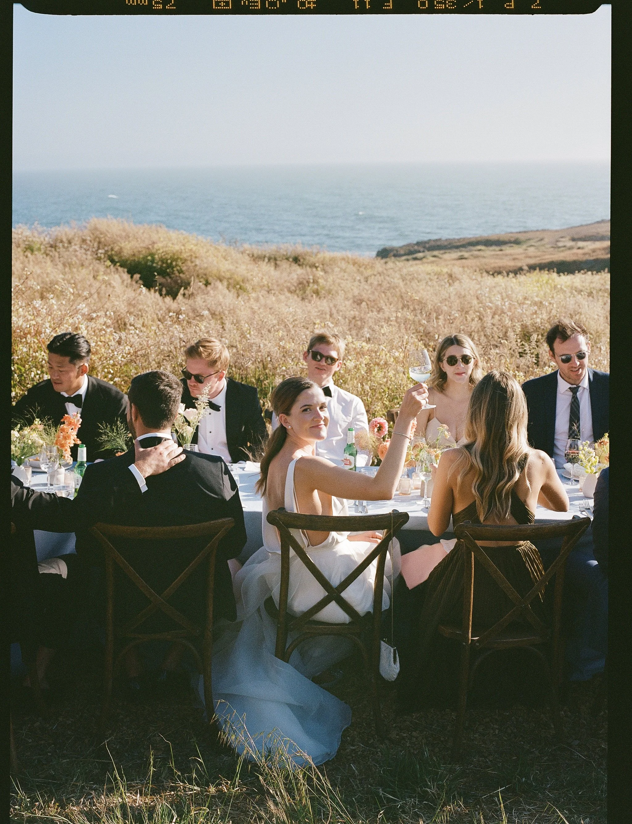 A group of people dressed in formal attire seated at a long outdoor table during their Sea Ranch wedding reception, with the ocean and coastline visible in the background under a clear sky.