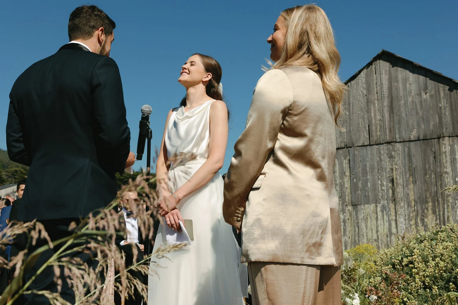 A wedding ceremony with a bride in a white dress and an officiant, a groom in a black suit, and a woman in a beige silk suit standing outdoors under a clear blue sky near a rustic wooden barn.