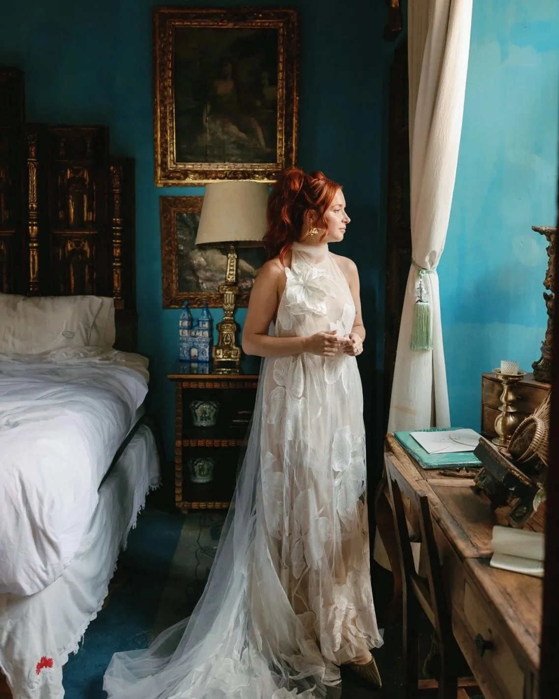 Red-haired woman in a flowing white gown standing beside a bed in an ornate blue bedroom at a historic wedding venue in san miguel de allende