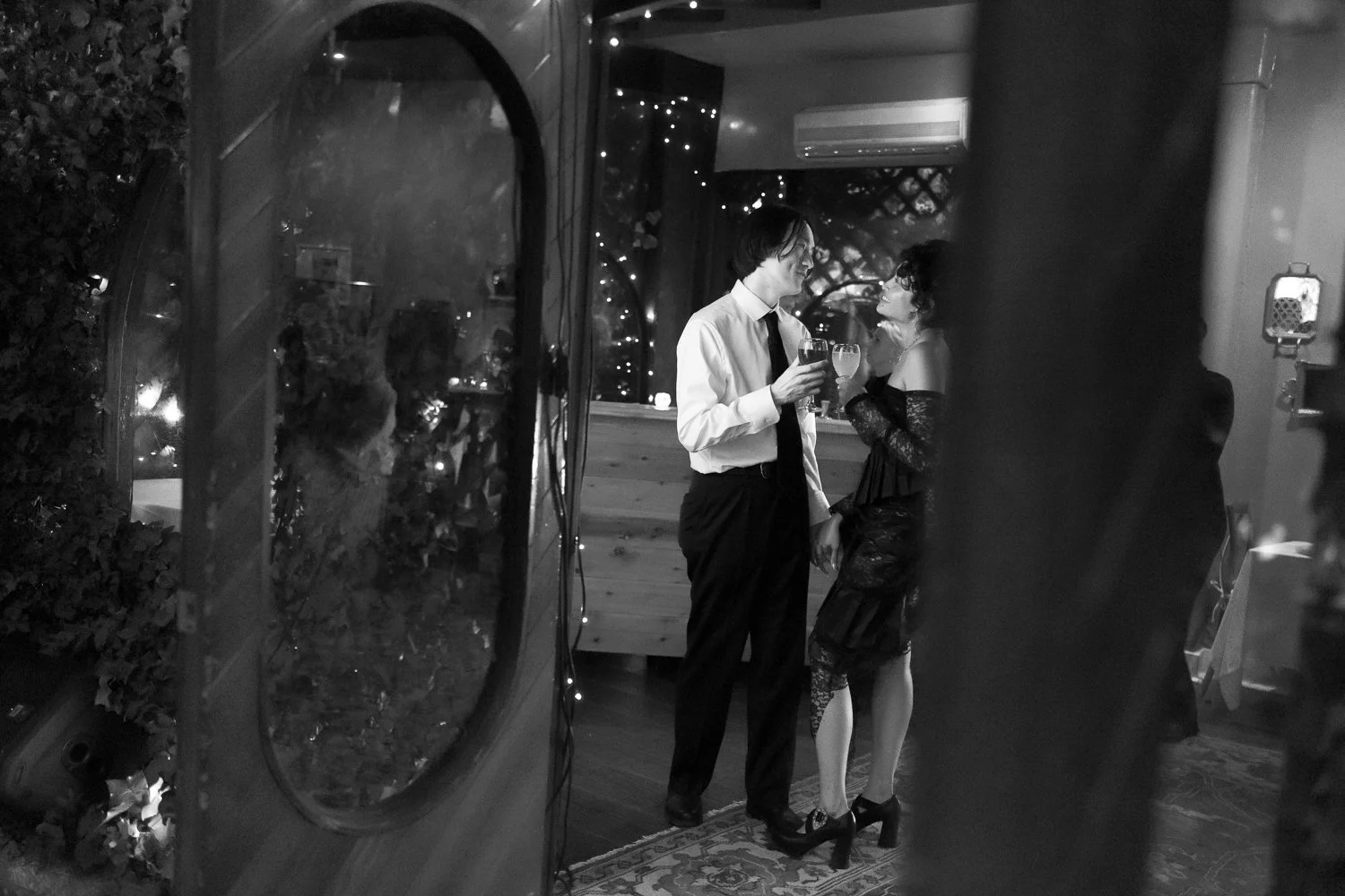 A black and white photo of a man and woman at a social gathering, possibly a party or wedding, standing close together and holding drinks, engaged in conversation. The man is dressed in a white shirt and dark trousers, while the woman is wearing a black lace dress with off-the-shoulder sleeves and high heels. They are indoors, with decorative lights visible in the background.