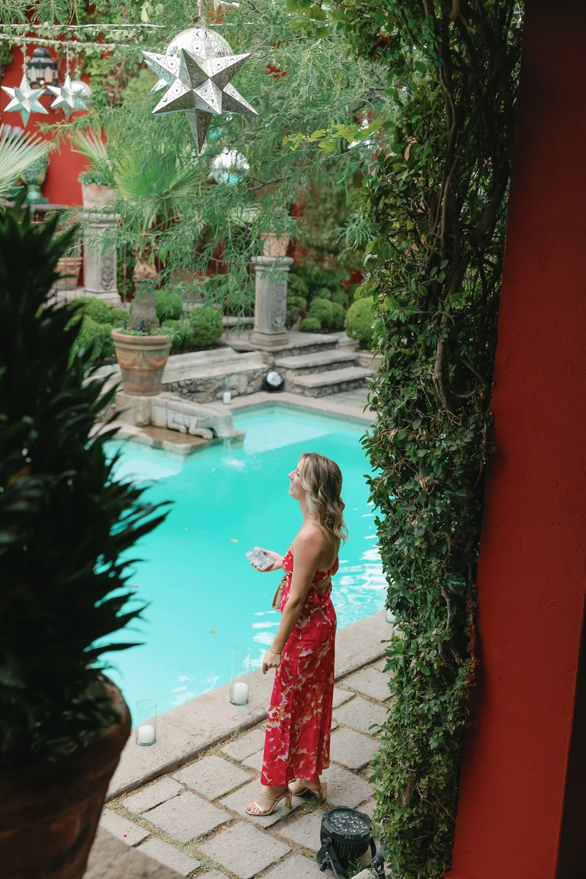 Woman in a red floral dress standing beside a turquoise courtyard pool surrounded by lush greenery and hanging star decorations