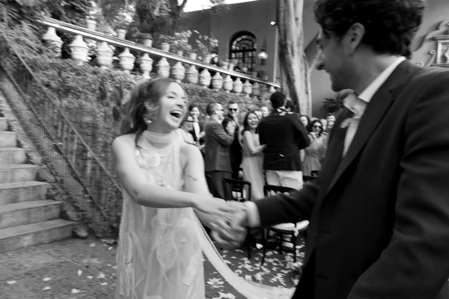 Black-and-white wedding reception scene with a smiling bride dancing with a groom outdoors as guests watch.