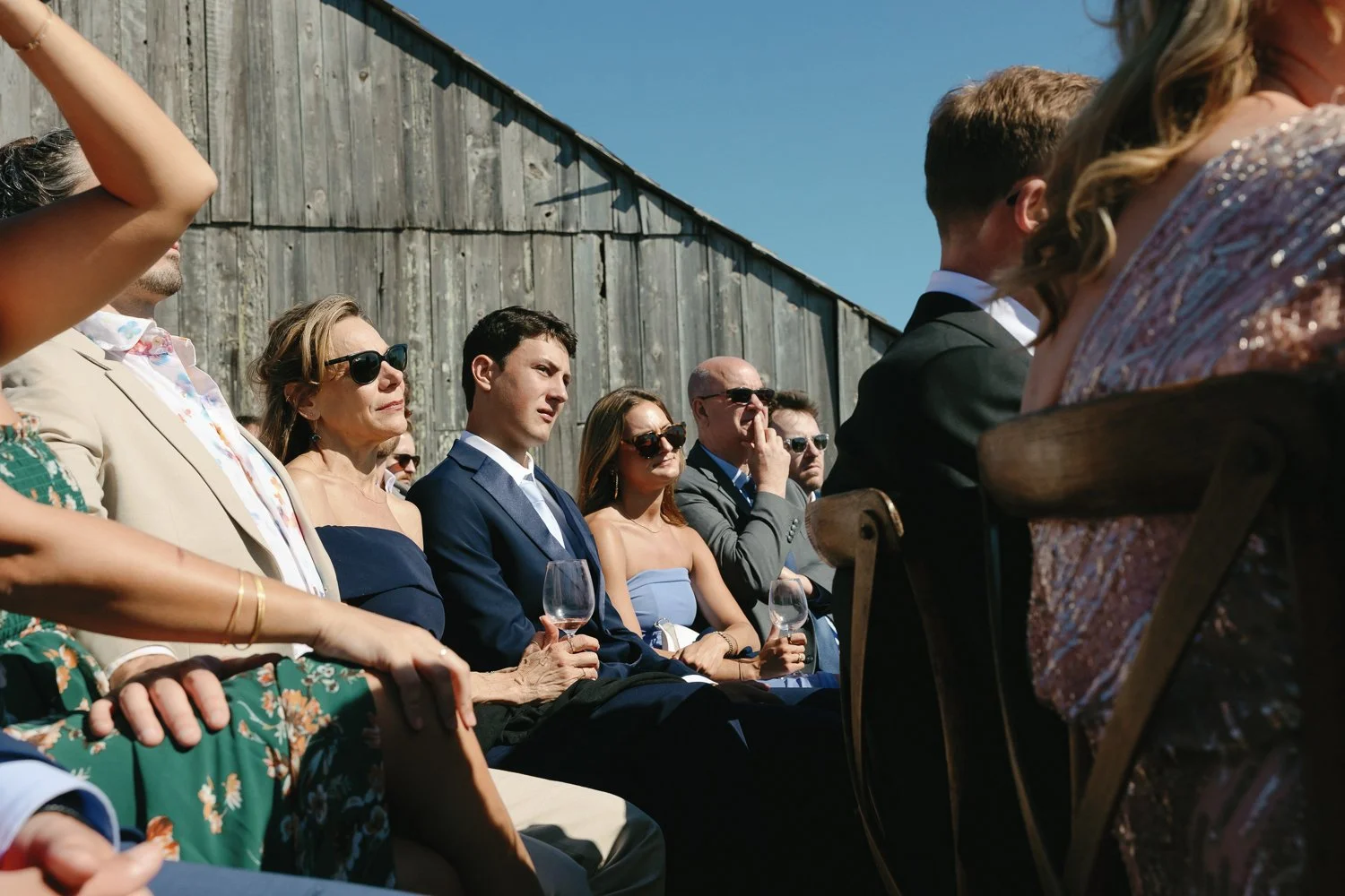 A group of people sitting outdoors at a formal event, some wearing sunglasses, with a weathered wooden wall in the background, under a clear blue sky.