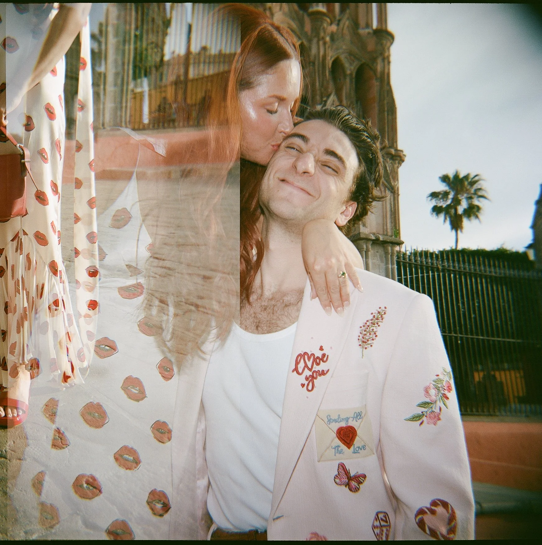 Red-haired woman kissing a smiling man in an embroidered pink and white blazer by PERTE D'EGO