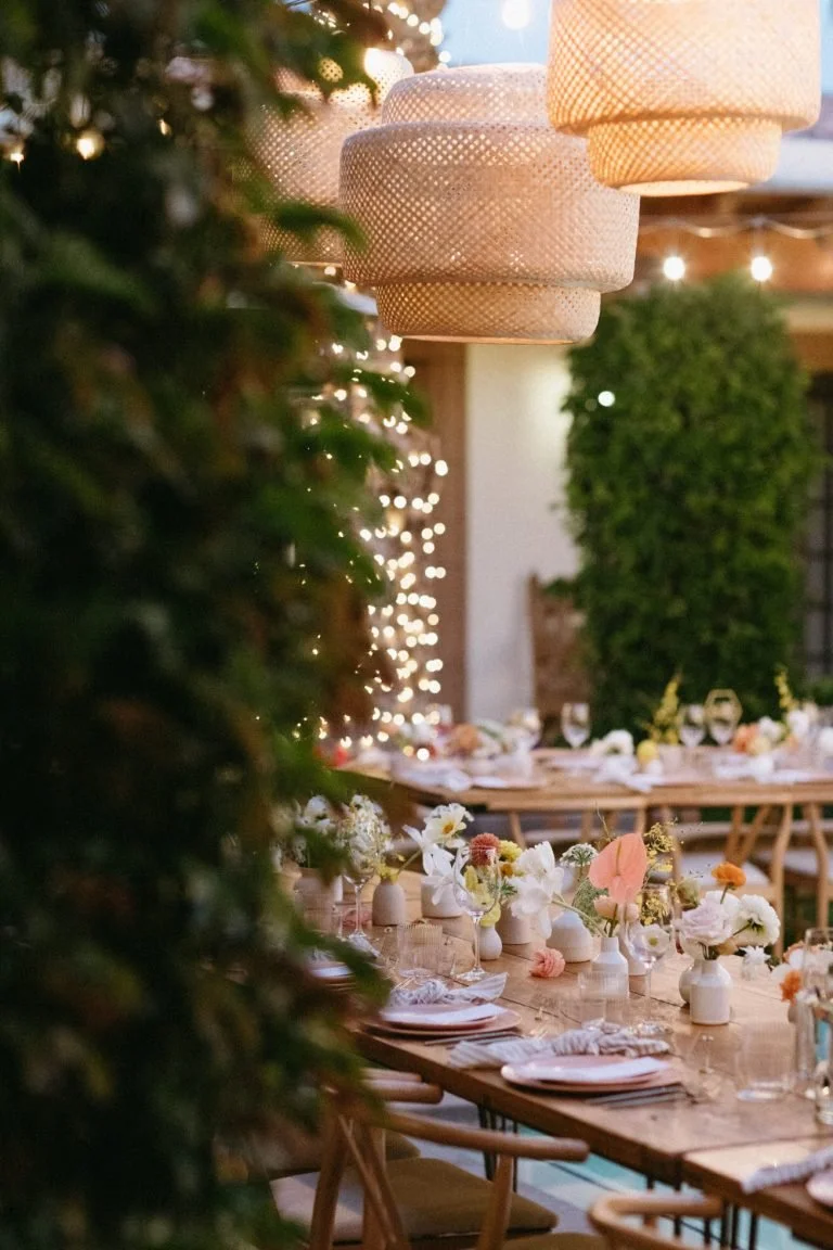 An reception around the pool at the bougainvillea estate decorated with hanging lanterns, string lights, and floral arrangements on a long wooden table.