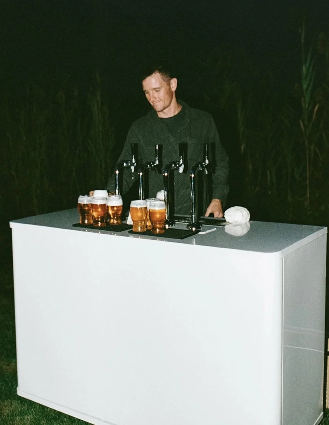 A man standing behind a white bar table with several glasses of beer on it at night.