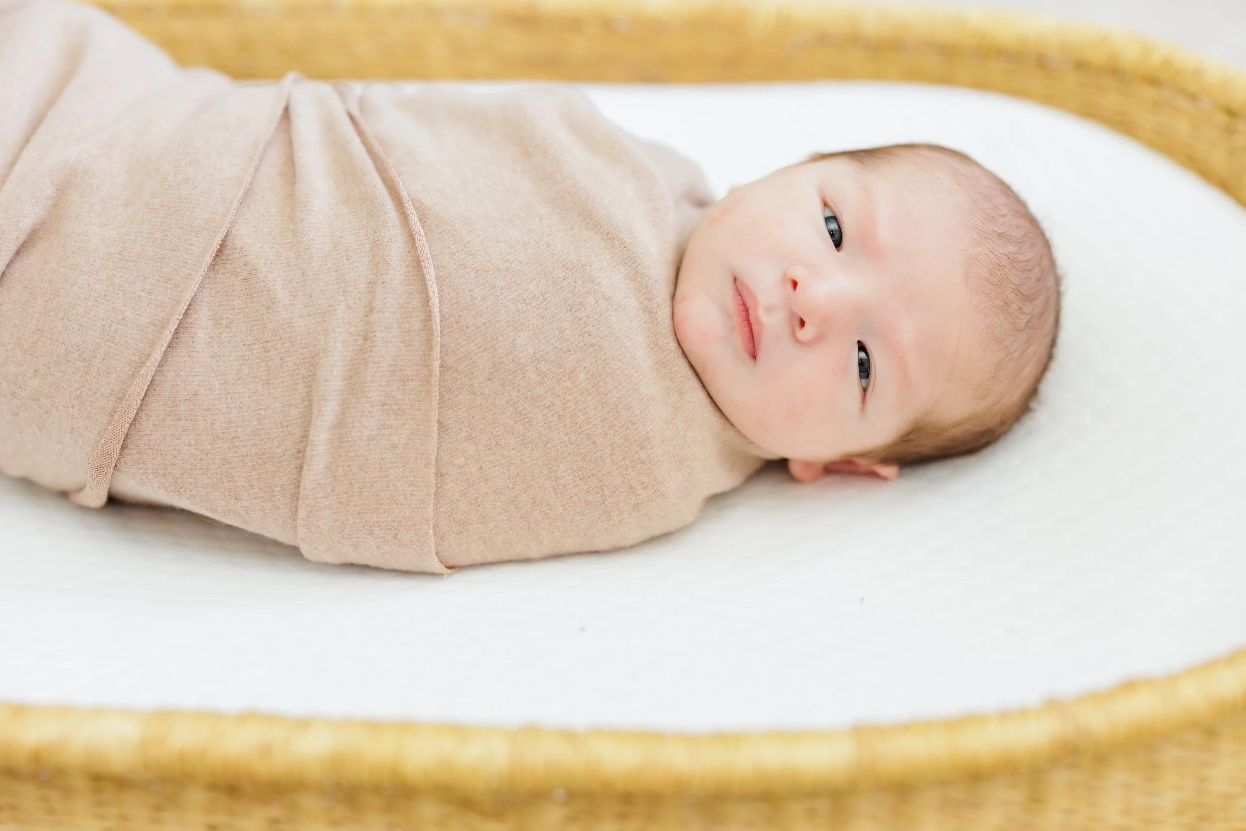 Close-up of a smiling baby wrapped in a beige blanket, lying in a wooden crib.