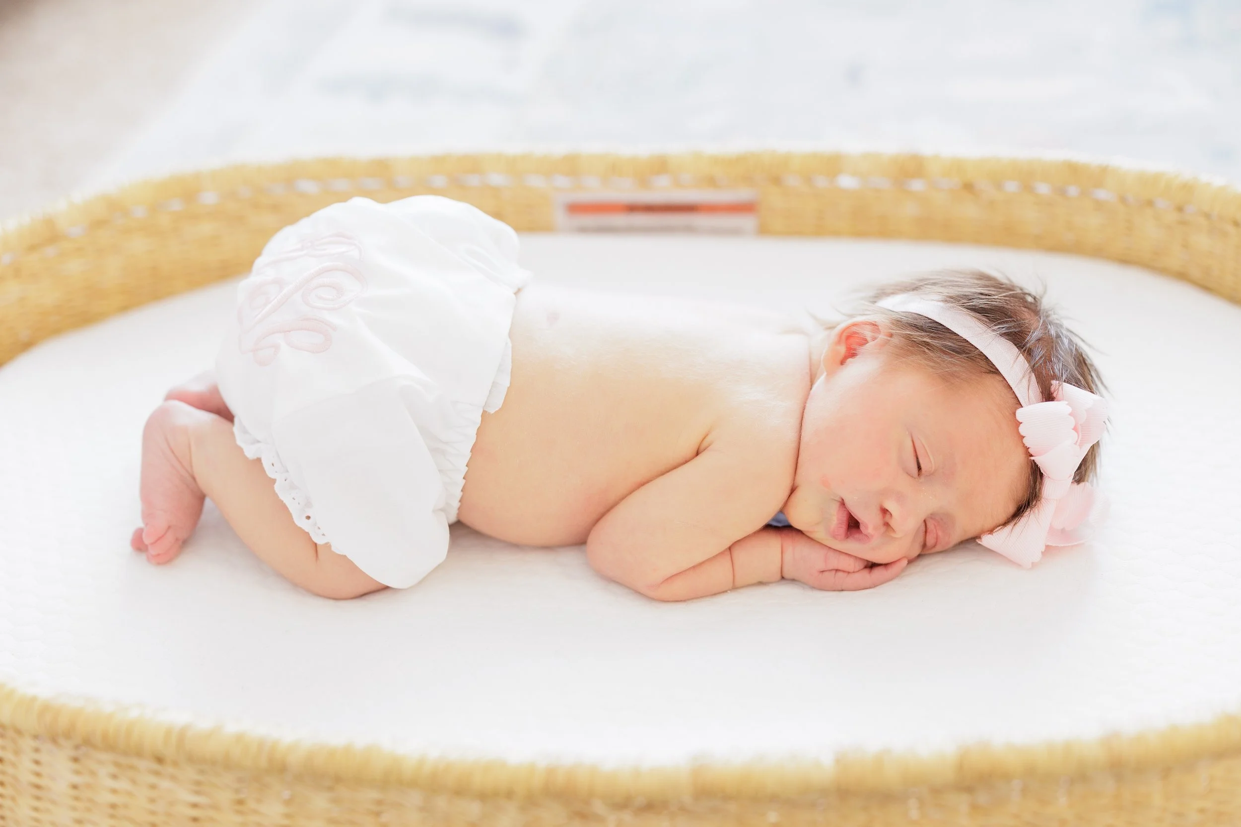 A baby is lying on its stomach inside a wicker basket, resting its head on folded arms with eyes closed, wearing a white headband with pink accents and a diaper.