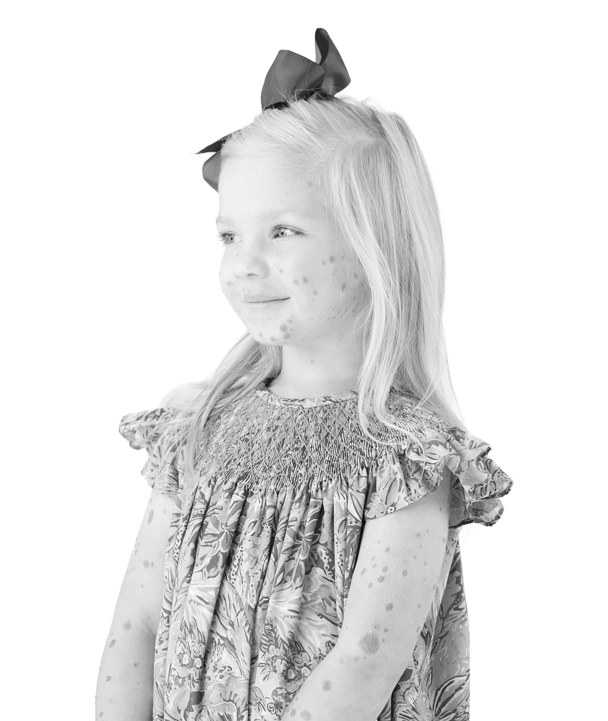 Black and white portrait of a young girl with long hair, wearing a patterned dress, looking to the side with a gentle smile, standing against a plain white background.