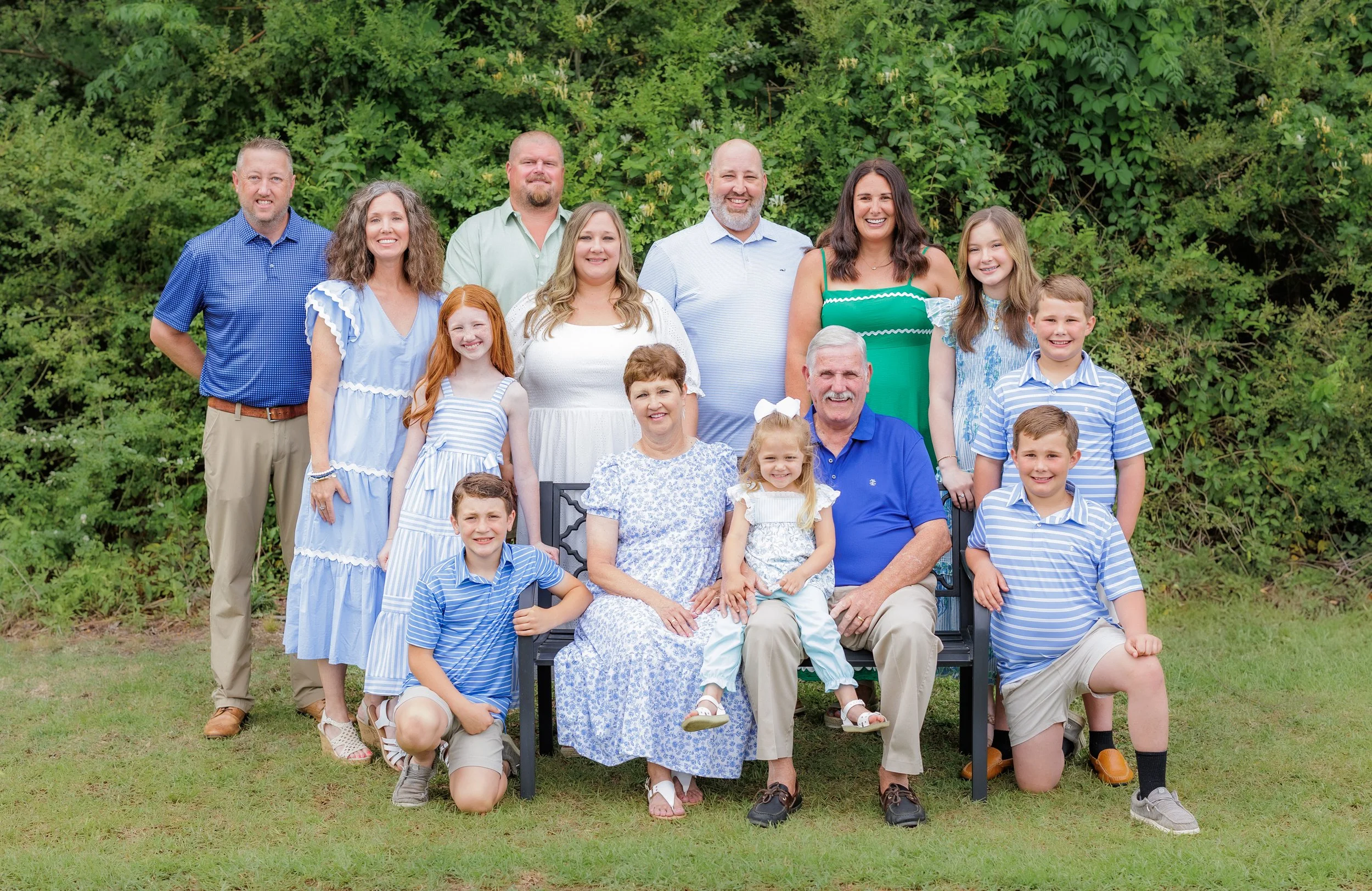 A large family group photo outdoors with green foliage background. The group includes older adults, middle-aged adults, teenagers, and children, all smiling and dressed casually, some wearing blue and white striped clothing.