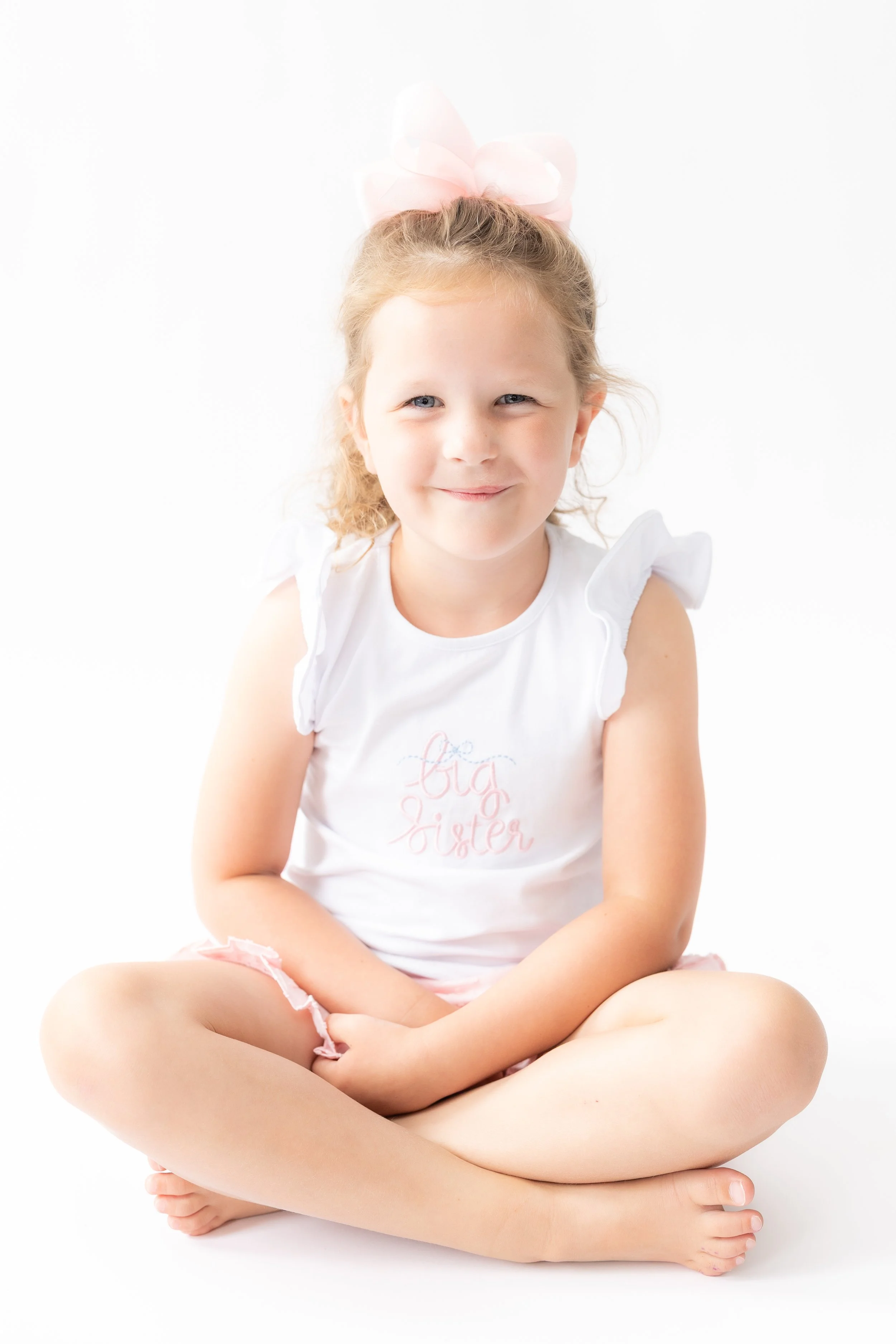 A young girl with blonde hair tied back with a pink bow, sitting cross-legged with arms resting on her knees, smiling gently against a plain white background. She is wearing a white sleeveless shirt that says 'Big Sister' and pink shorts.