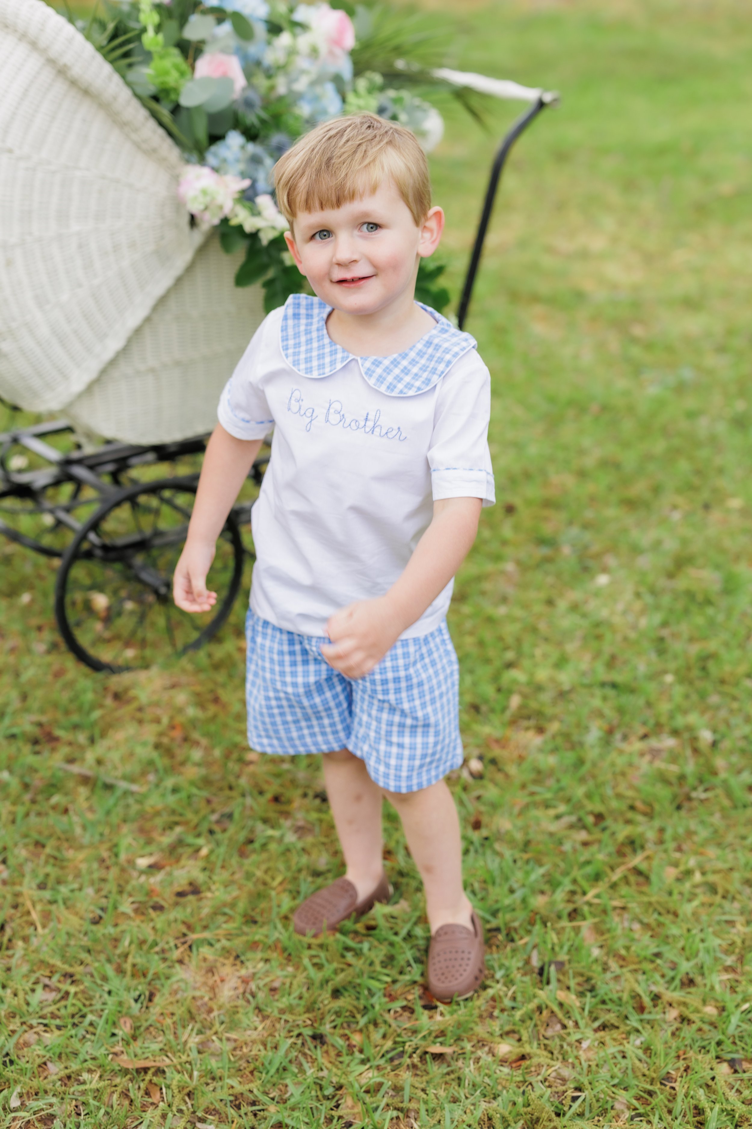 A young boy with red hair and blue eyes standing outdoors on grass, wearing a white shirt with a blue gingham collar and text that reads 'Big Brother,' blue gingham shorts, and brown slip-on shoes. A vintage white wicker stroller filled with colorful