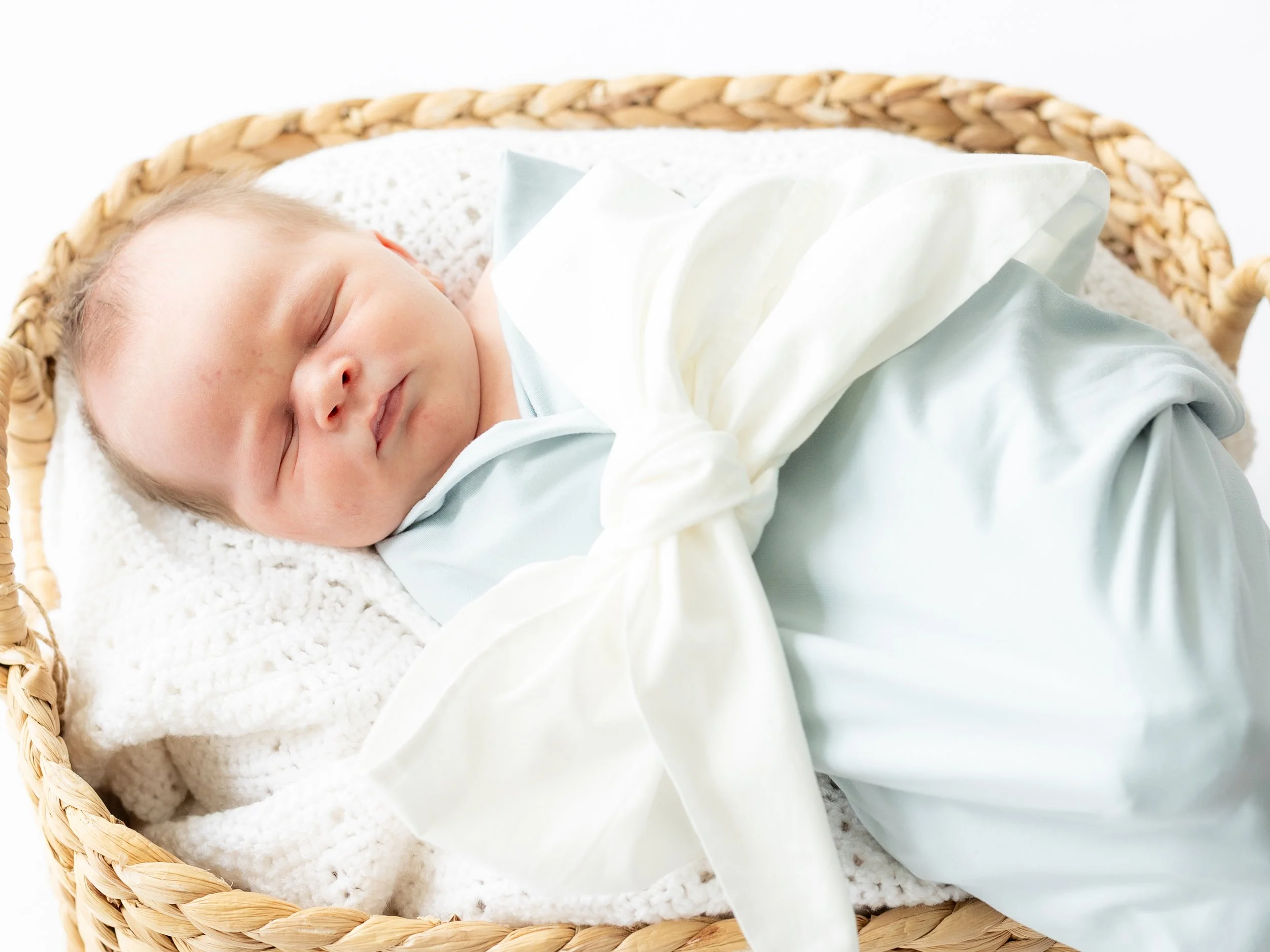 Newborn baby sleeping in a woven basket with soft white bedding and a white blanket.