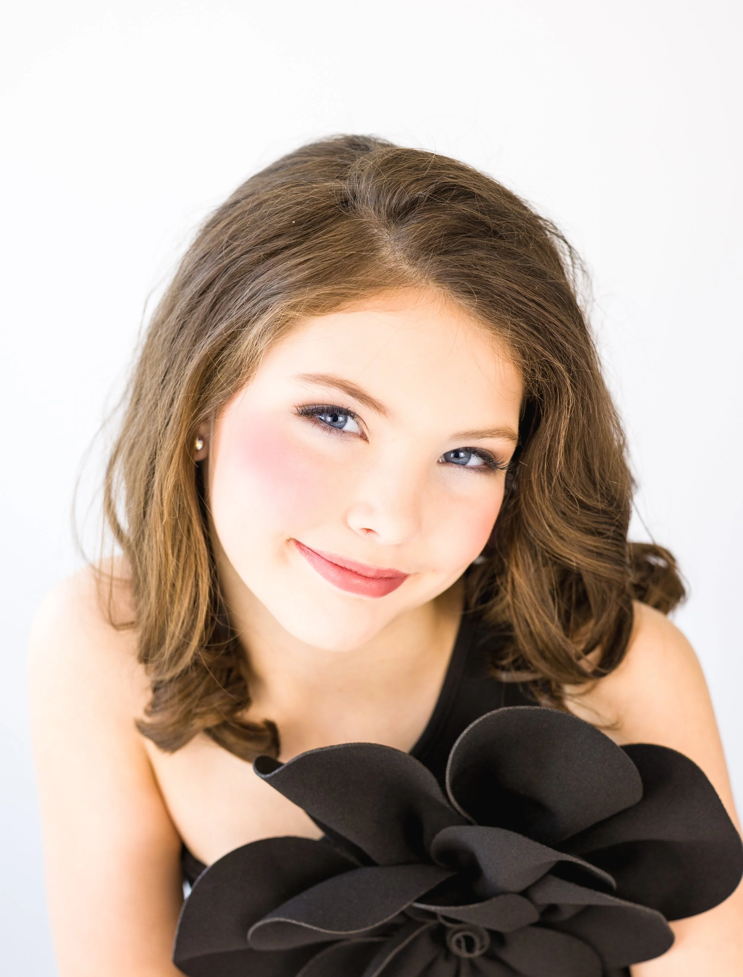 A woman with brown hair styled in loose waves, blue eyes, and makeup smiling at the camera, wearing a black top and a large black fabric flower necklace.