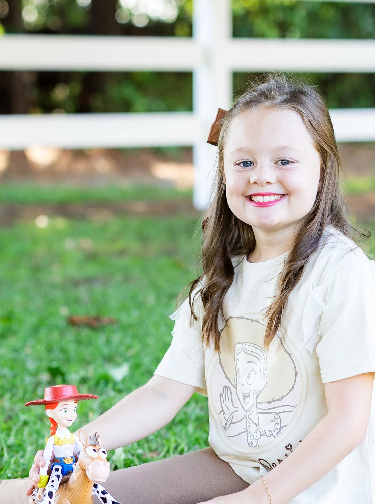 A young girl with long brown hair, wearing a beige T-shirt with a cartoon skeleton on it, smiling while playing with Woody and Bullseye toys outdoors on grass.