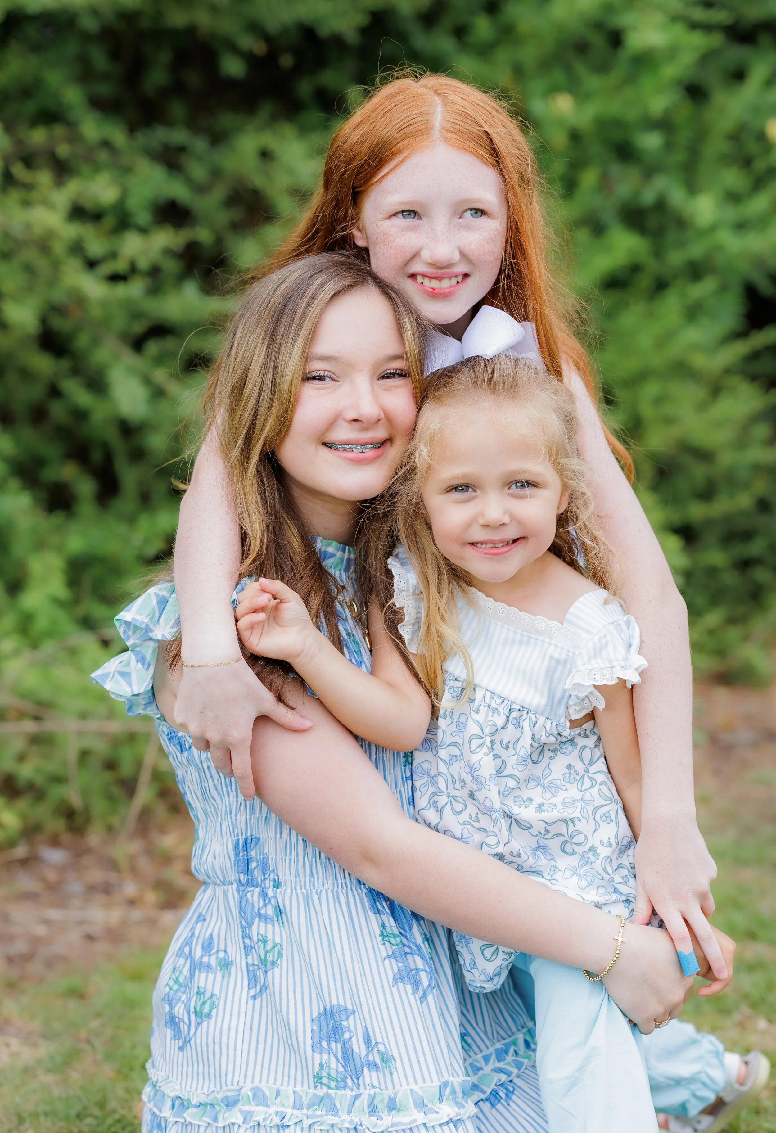 Four young girls with red hair and blonde hair, smiling and posing outdoors with greenery in the background.