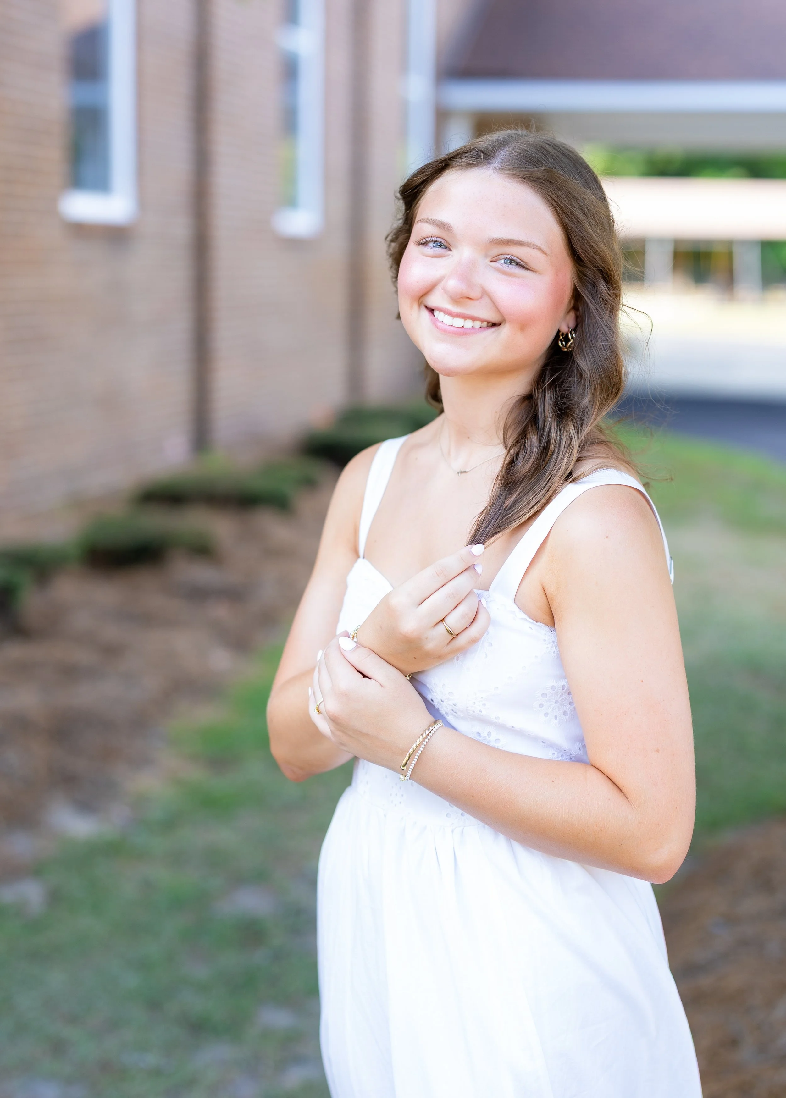 A young woman with blue eyes and wavy brown hair in a white dress standing outdoors near a brick building, smiling and holding her hands together near her chest.