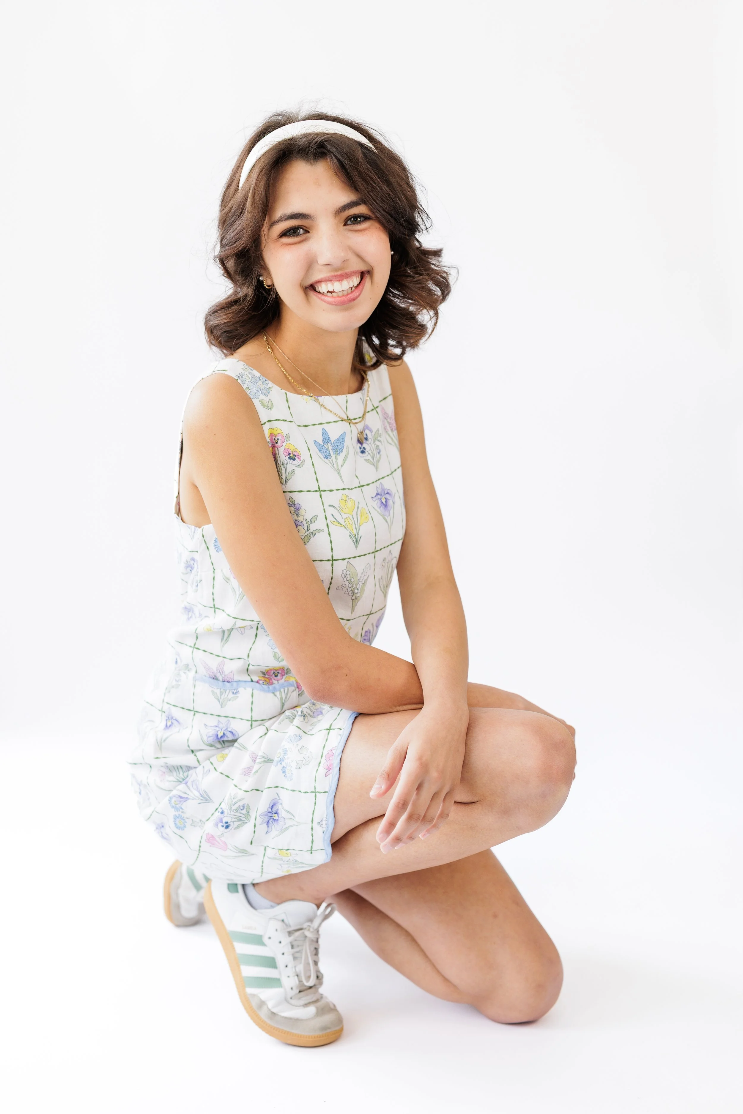A young woman with dark wavy hair wearing a headband, floral sleeveless top, and sneakers, sitting on one knee and smiling against a white background.