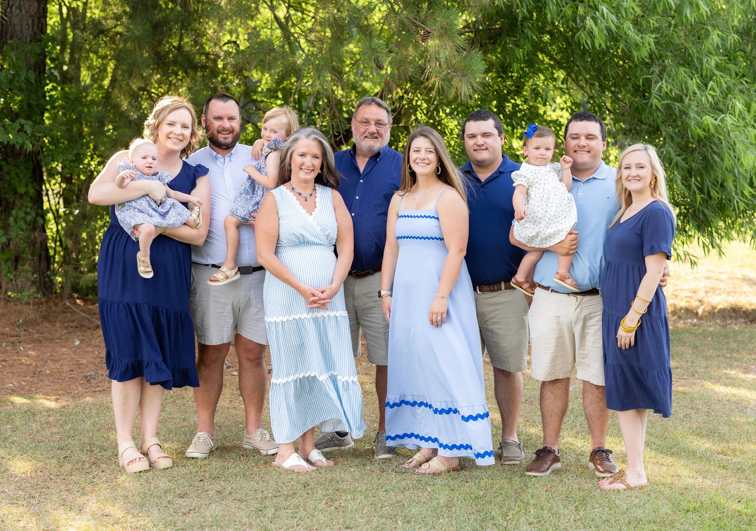 A large family group standing outdoors on grass, with green trees in the background, smiling and posing for a photo.