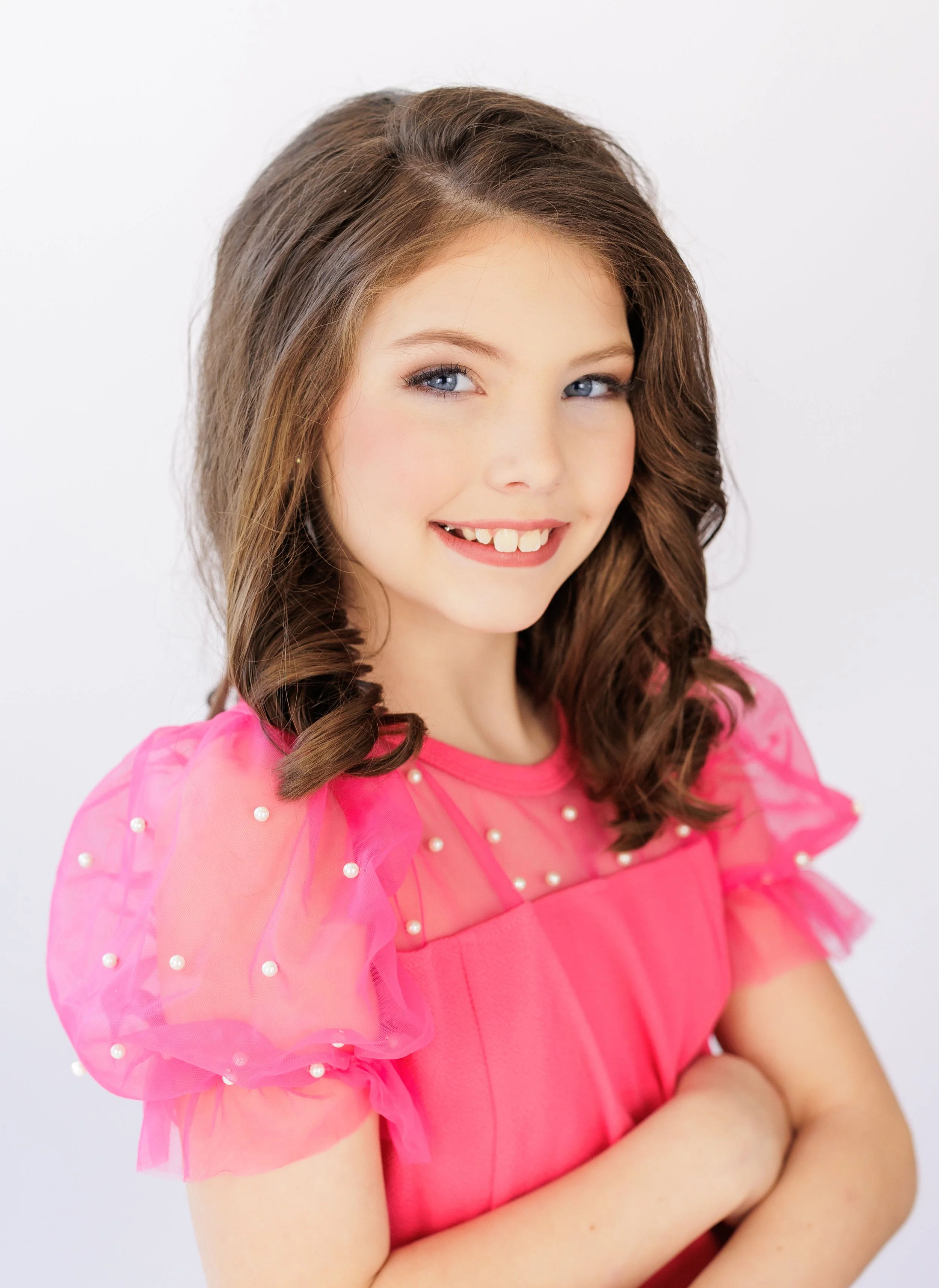 A young girl with long brown hair and blue eyes, smiling and wearing a pink dress with sheer, puffed sleeves adorned with pearls.