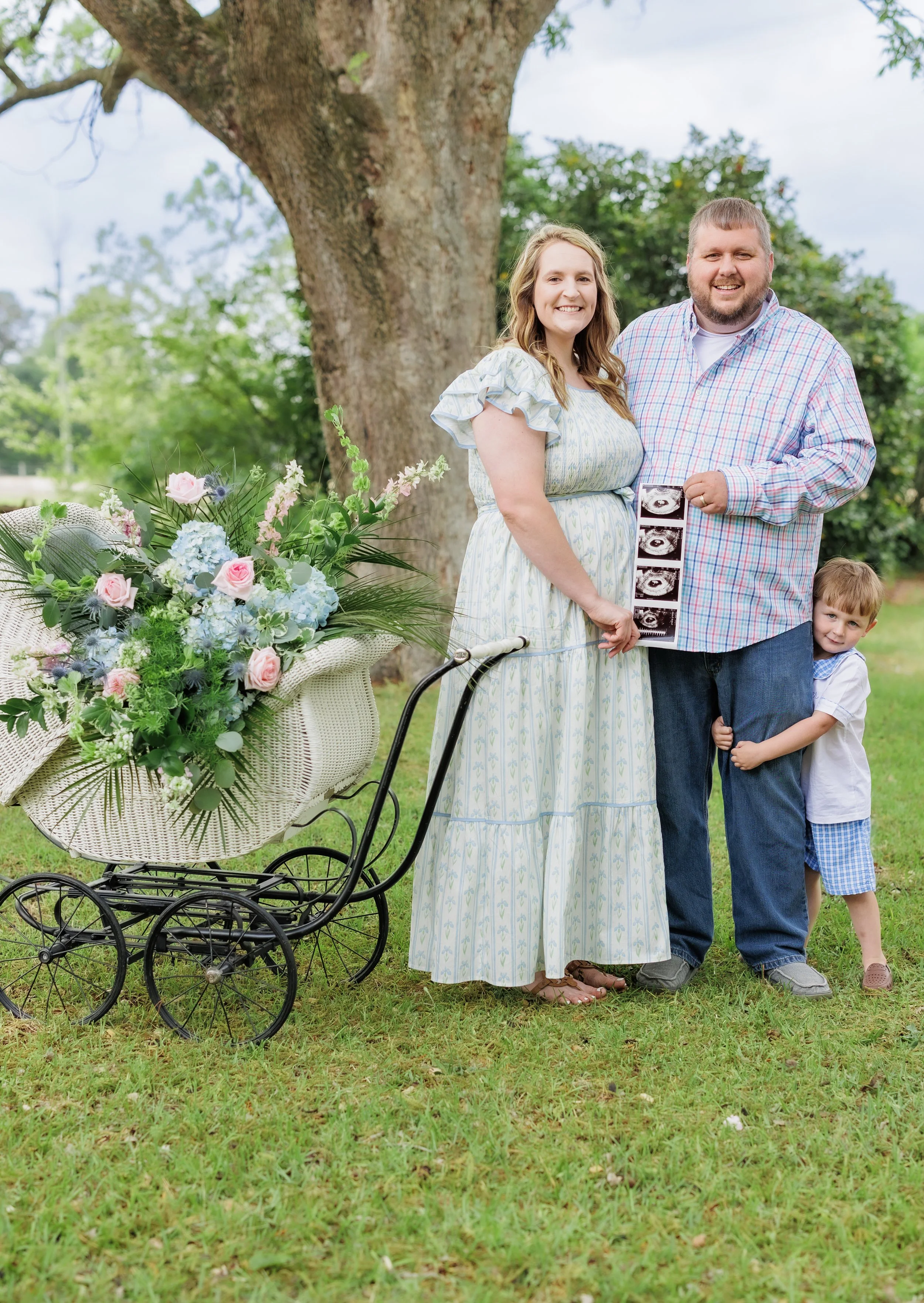 A couple with a young boy standing outdoors next to a basket of flowers on a bicycle, holding ultrasound images, smiling in front of a large tree.