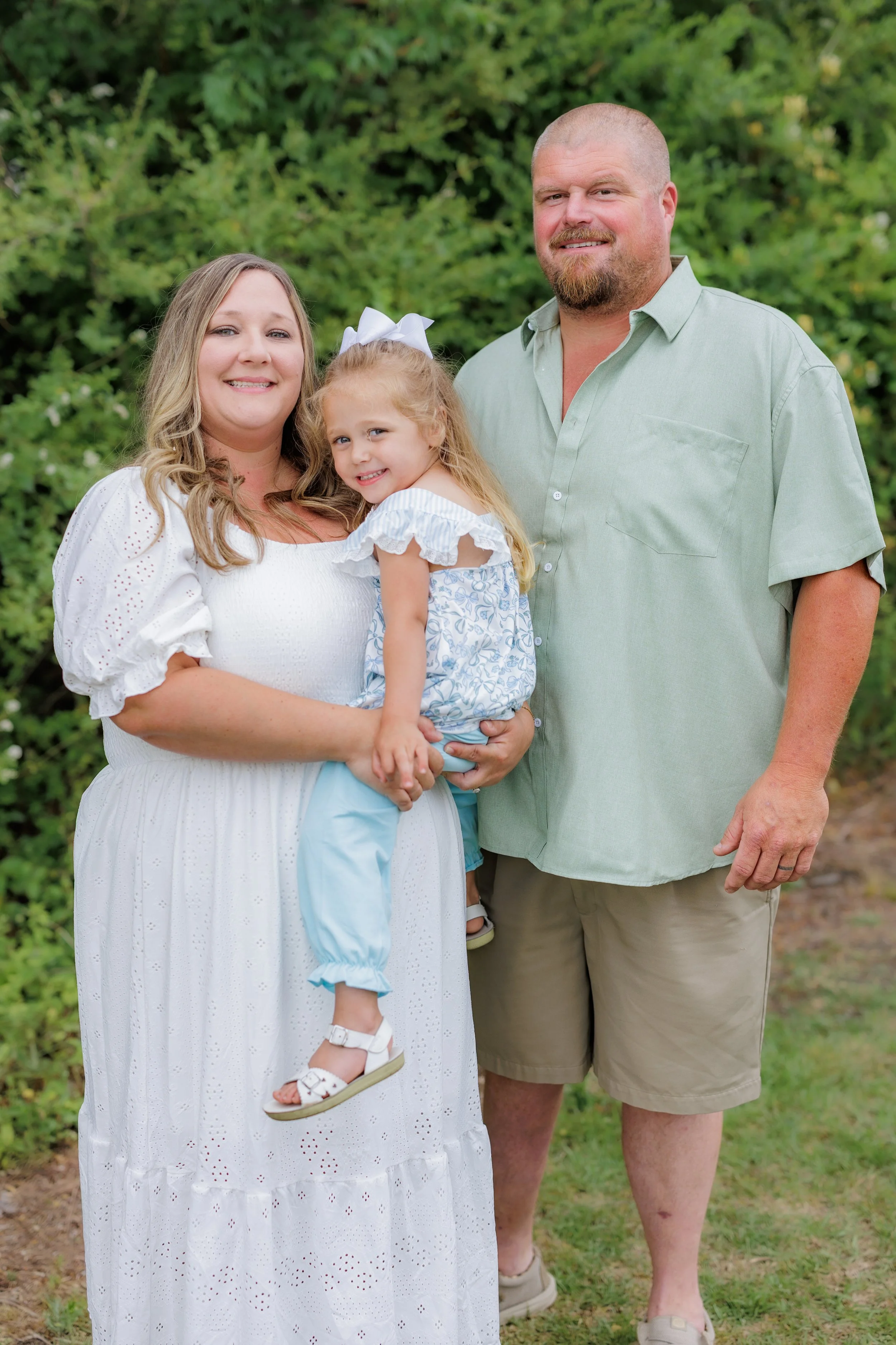 A family of three outdoors. A woman in a white dress holding a young girl dressed in blue, standing next to a man in a light green shirt and khaki shorts. They are in front of green bushes.