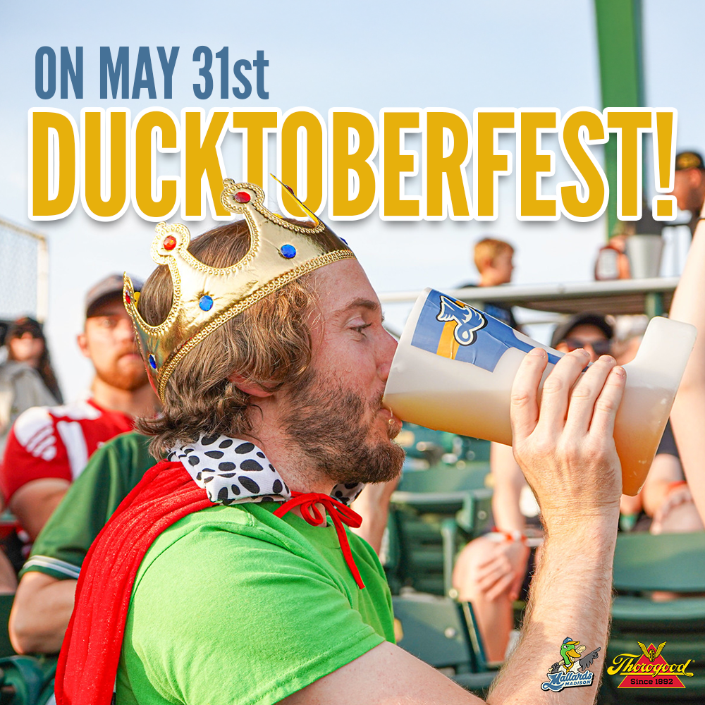 A man wearing a gold crown, green shirt, and red cape drinking beer at a sports stadium during DuckToberfest event on May 31st, with other fans in the background.