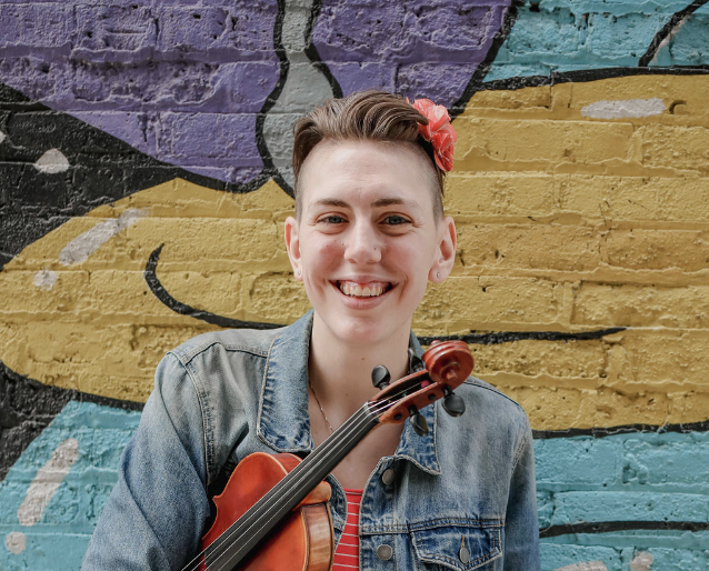 Miss Grube smiling while holding a violin in front of colorful graffiti wall.